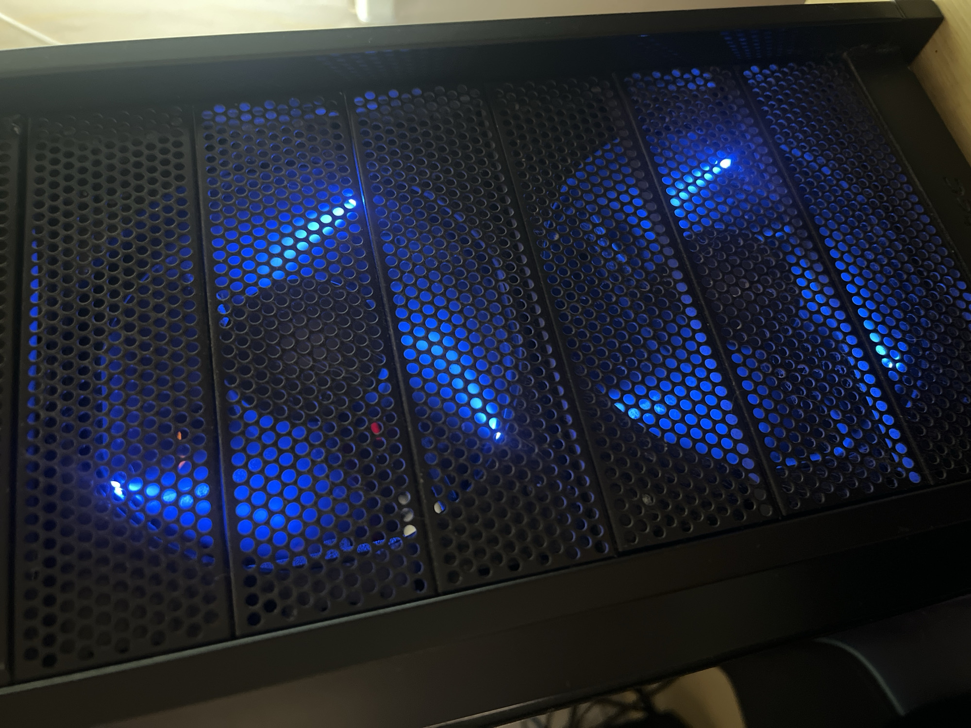 The image shows a close-up, eye-level, full shot of a black computer tower with multiple levels of perforated metal mesh. Behind the mesh, there are fans with bright blue LED lights, illuminating the interior of the case through the circular perforations. The lighting suggests the computer is powered on, and the blue lights create a futuristic and technical aesthetic. The background is blurred, with hints of a desk surface and a white wall or object. The image appears to be taken indoors in an indoor setting, possibly a home or office. There are no people or animals visible in the image.
