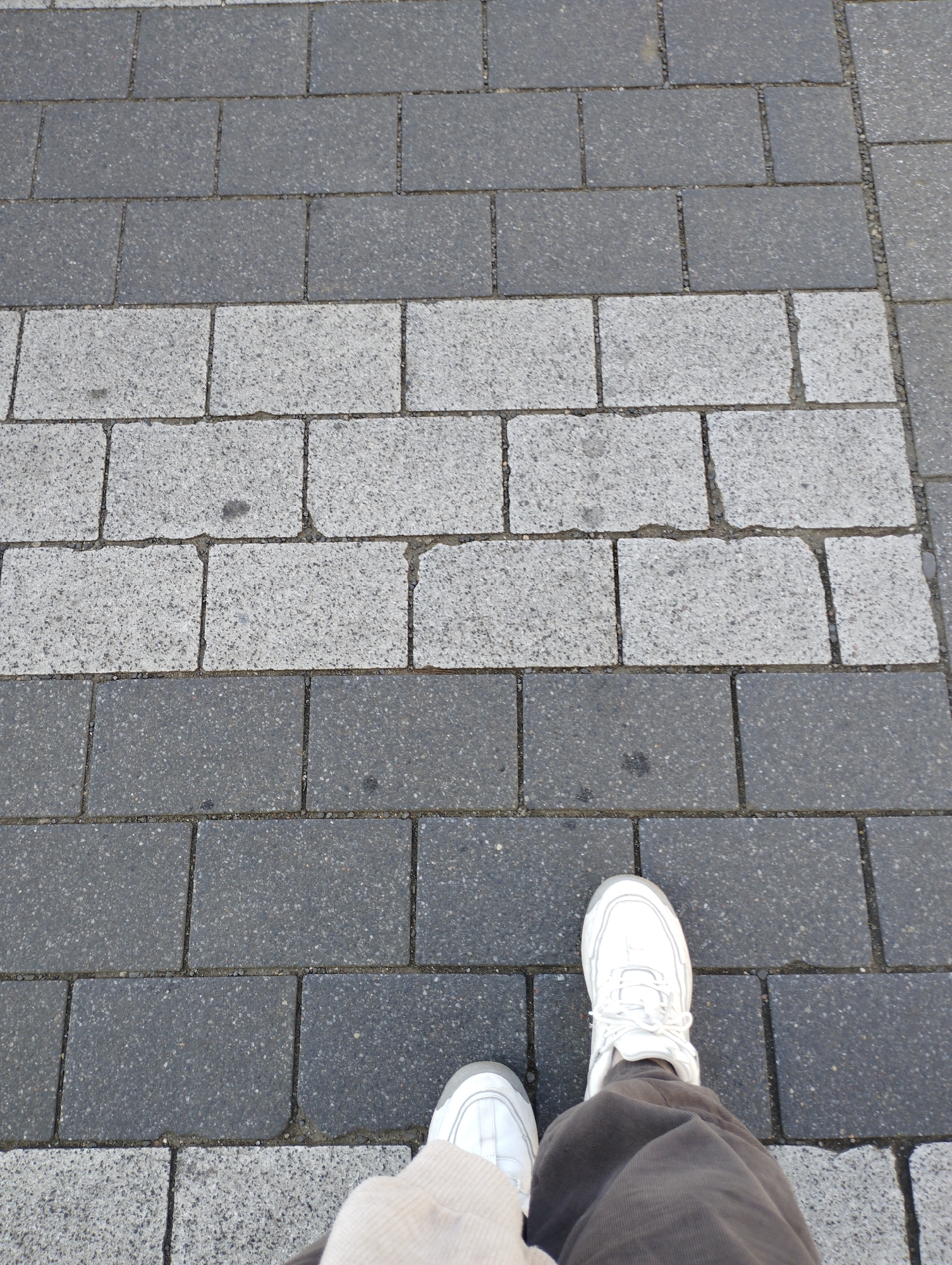 This is a first-person perspective, ground-level photograph taken on a sidewalk composed of rectangular paving stones. The sidewalk features a distinct pattern of alternating bands of dark grey and lighter grey stones. In the lower portion of the image, a person's legs and feet are visible, wearing white athletic shoes and brown trousers. The person is standing on the sidewalk, facing downwards. The environment appears to be an urban setting, likely a pedestrian area. The image was taken in Szczecin, Poland.