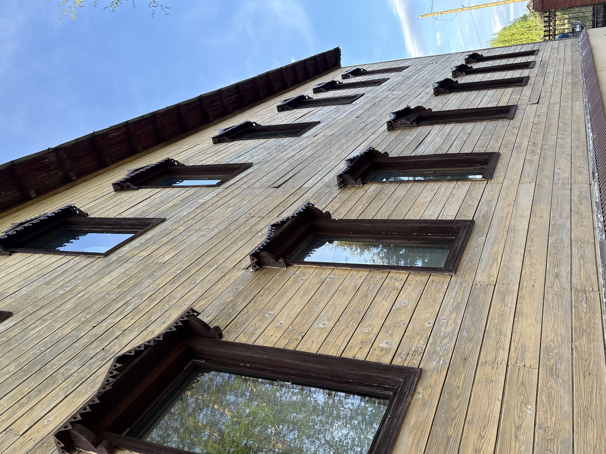 The image depicts a building exterior in Zavod Mikhailovskii, Russia, notable for its distinct architectural features. The facade is constructed from vertical wooden planks, exhibiting a light yellow hue with signs of weathering. A series of windows are arranged in a vertical pattern along the facade, each framed with dark brown trim. The windows are adorned with decorative, sawtooth-edged trim along the top. The angle of the shot emphasizes the verticality of the building, with the sky visible in the background. The environment appears to be a residential or mixed-use area, as indicated by the presence of trees reflected in the windows.