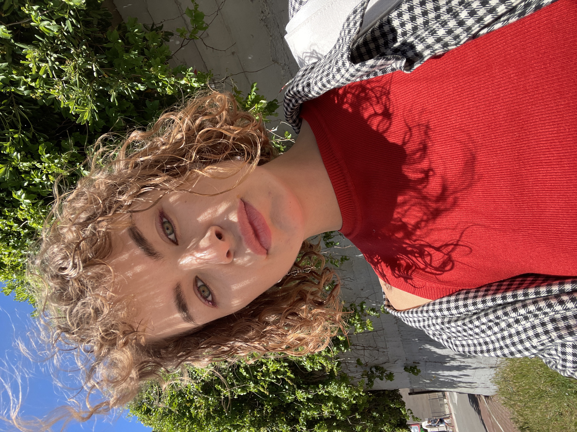 A woman with curly, light brown hair, fair skin, and green eyes is positioned diagonally in the frame, wearing a red top and a black and white patterned jacket draped over her shoulder. She is standing outdoors, with green foliage and the corner of a building visible behind her. The blue sky is visible in the upper left corner. The location is identified as Rafailovići, Montenegro.