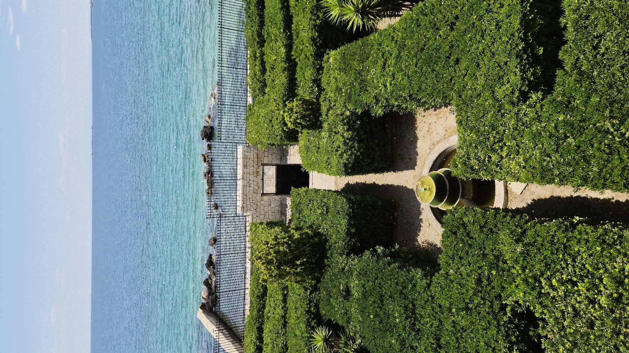 An aerial view captures a well-maintained garden in Taranto, Italy, overlooking the Ionian Sea. The garden features neatly trimmed green hedges forming a maze-like structure, with gravel pathways winding through. In the center of the garden, a tiered fountain with green water sits within a circular basin. Beyond the hedges, a stone wall with an arched gateway leads to a darker, enclosed space. A black metal fence separates the garden from the sea, with a concrete walkway and rocks visible along the shoreline. The sea itself is a vibrant turquoise, stretching to the horizon under a clear blue sky. There are no people or animals present in the image.