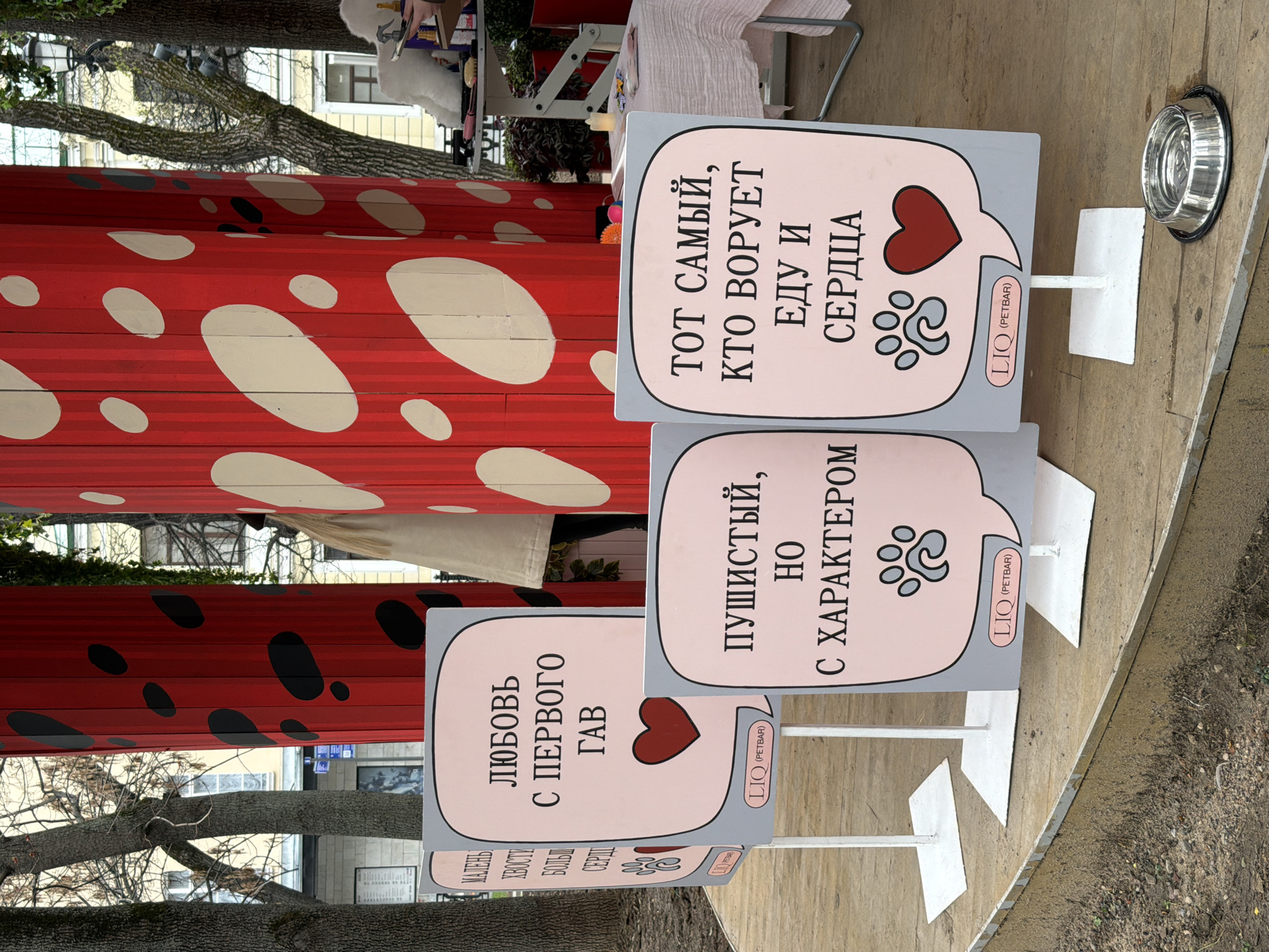 This outdoor photograph captures a scene likely from a pet-themed event or market in Moscow, Russia. The primary subjects are four freestanding, speech-bubble-shaped signs with affectionate and humorous messages about pets. The signs feature a light pink background, dark gray borders, and include a red heart and/or paw print graphics. They are supported by white metal stands. A metal pet bowl is visible on the ground to the far right, reinforcing the pet theme.
In the background, red structures with white and black spots, resembling large mushrooms or ladybugs, serve as decorative elements or part of stalls. Bare tree branches suggest the season is late autumn, winter, or early spring. Faint architectural details of buildings are visible through the trees. The ground is paved, indicating an urban or park setting.
The visible text on the signs (in Cyrillic) translates to:
- "LOVE FROM THE FIRST WOOF" (with a heart and paw prints)
- "FLUFFY, BUT WITH CHARACTER" (with paw prints)
- "THE ONE WHO STEALS FOOD AND HEARTS" (with a heart and paw prints)
A partially visible leftmost sign shows "SMALL TAILS BIG HEART." All signs include the branding "LIO (PETBAR)," indicating a sponsor or brand related to pets. The overall atmosphere conveyed by the signs is warm, lighthearted, and appreciative of animals.
