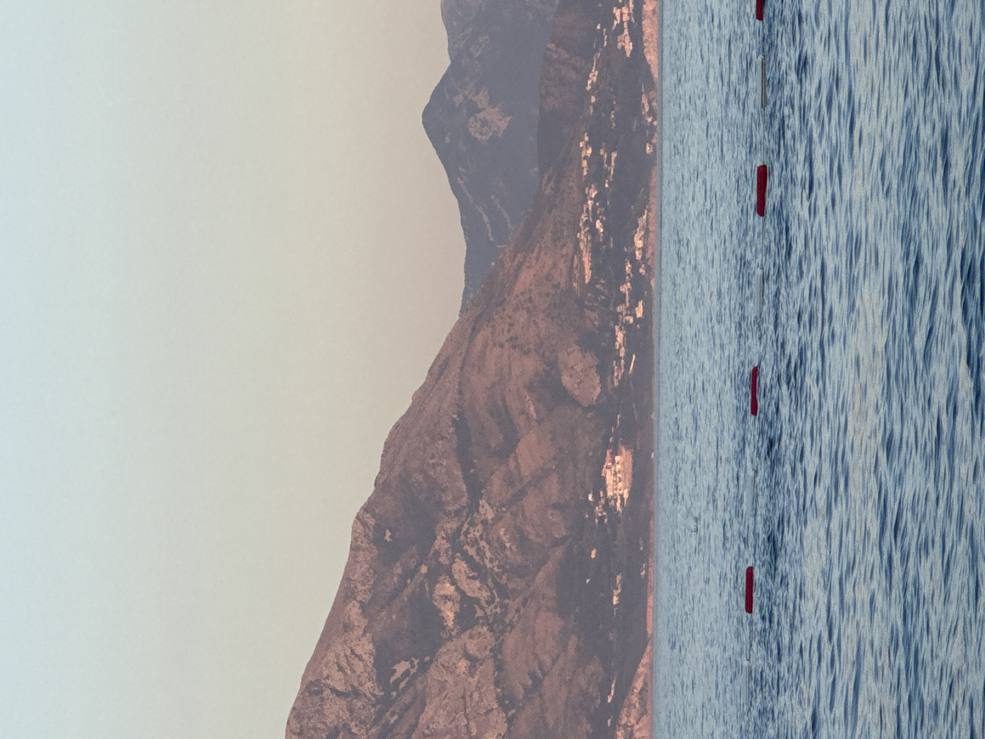 A horizontal, medium shot captures a serene seascape under a pale, gradient sky, transitioning from light blue at the top to a soft peach hue near the horizon. The foreground is dominated by a body of tranquil, blue water, marked by subtle ripples and a series of three red, rectangular buoys spaced evenly across the frame. In the mid-ground, a densely vegetated hillside is visible, dotted with small, light-colored buildings, suggesting a coastal settlement. The background features a majestic mountain range, its rocky slopes rendered in muted tones of brown and grey, with shadowed crevices and lighter, sunlit peaks. The overall atmosphere is peaceful and idyllic, with a clear division between the calm water, the inhabited hillside, and the imposing mountains. The scene is consistent with the coastal geography of Prijevor, Montenegro, known for its proximity to the Adriatic Sea and its mountainous terrain.