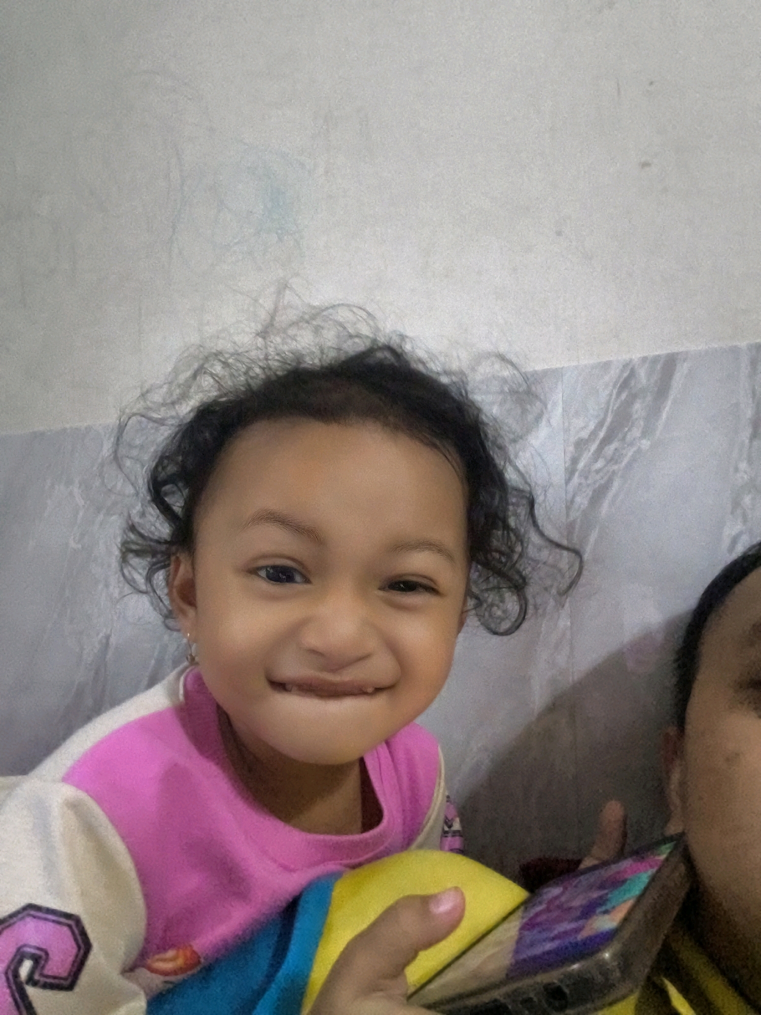 A young child, appearing to be of East Asian or Southeast Asian descent, with dark curly hair, is captured in a close-up selfie. The child is smiling and wearing a multi-colored shirt with pink and white sections. The background consists of a wall with a textured pattern and what appears to be a folded sheet or curtain. Part of another person's face and a smartphone are visible in the foreground. The image quality is moderate with some graininess. The context of the location in Karangtalun, Indonesia, is not discernible from the visual information in the image itself.