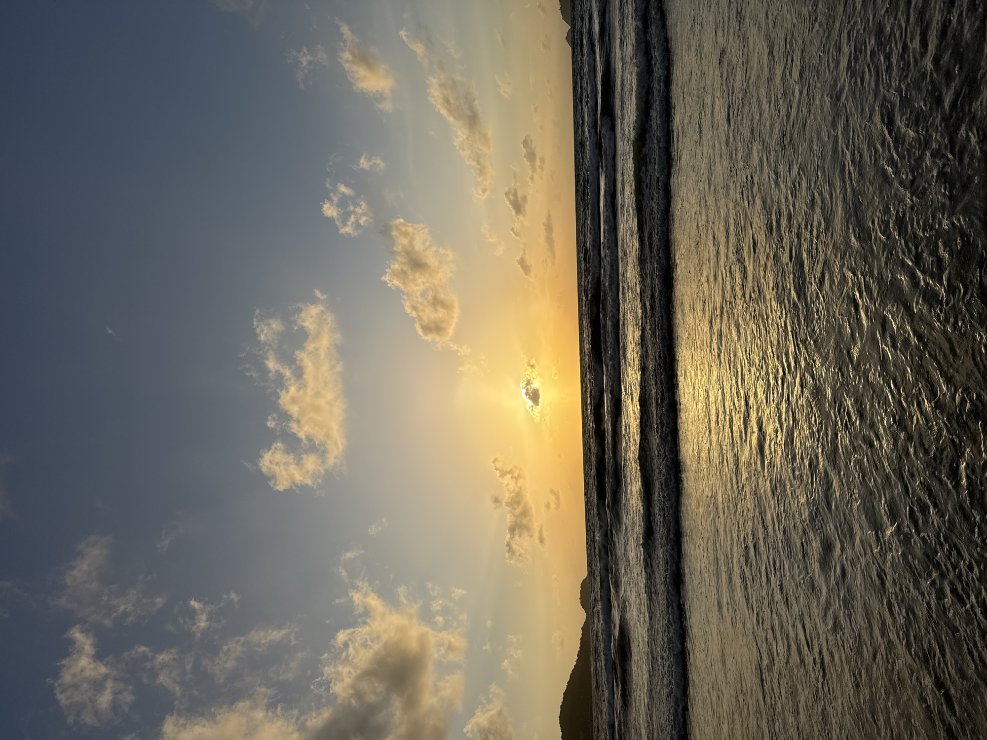 This is a photograph of the beach in Patong, Thailand, at sunset. The sky is a gradient of blue and orange, with scattered clouds. The sun is low on the horizon, casting a golden glow over the scene. The ocean is calm, with gentle waves rolling towards the shore. There are dark silhouettes of hills or land masses on either side of the beach. The water in the foreground reflects the warm colors of the sunset. No people are visible in the image.