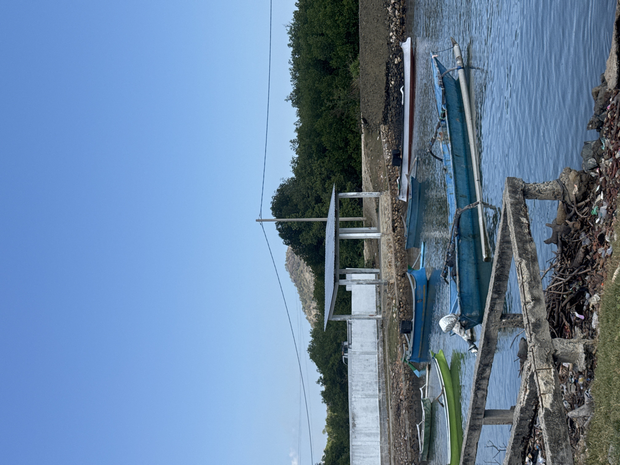 The image shows a body of water with several boats. In the background, there is a white wall, a covered structure with a metal roof supported by pillars, and lush green trees. Beyond the trees, a hilly landscape is visible under a clear blue sky. The foreground features a concrete structure with exposed rebar, debris, and dry vegetation. The water is calm and reflects the surrounding environment. There are no people visible in the image. The scene appears to be a coastal or riverine area in Kertasari, Indonesia, likely a small dock or mooring area.