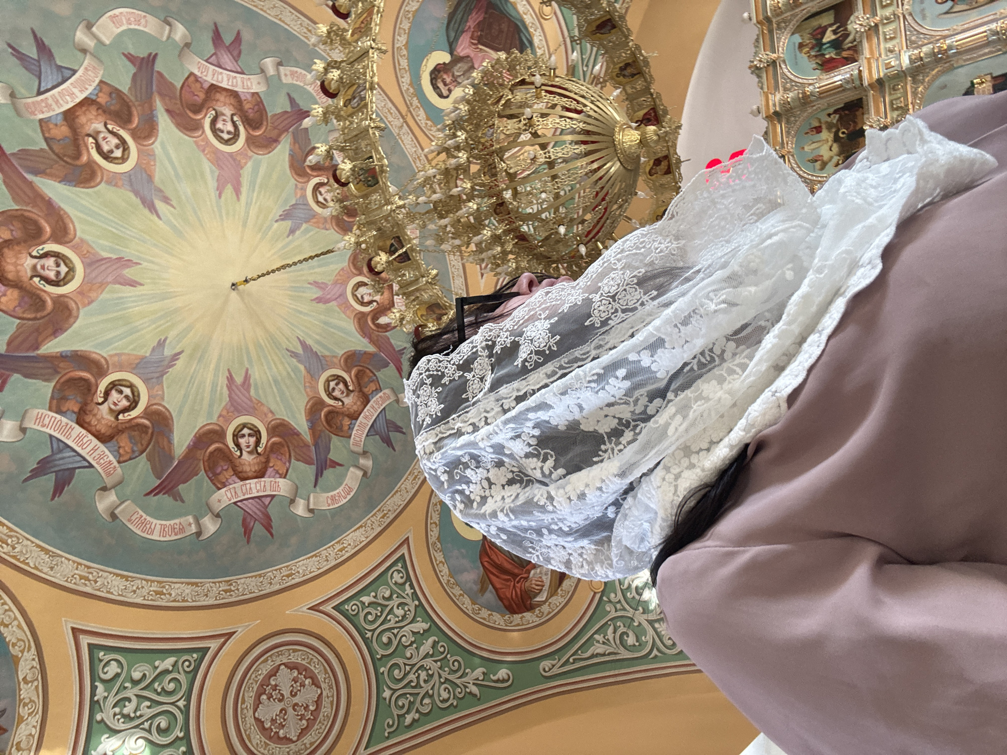 The interior of a church is visible in the image. An ornate chandelier hangs from a ceiling painted with a circular arrangement of angels. A woman in a mauve jacket and a white lace head covering is standing in the foreground. The city and precise location of the church are unknown.