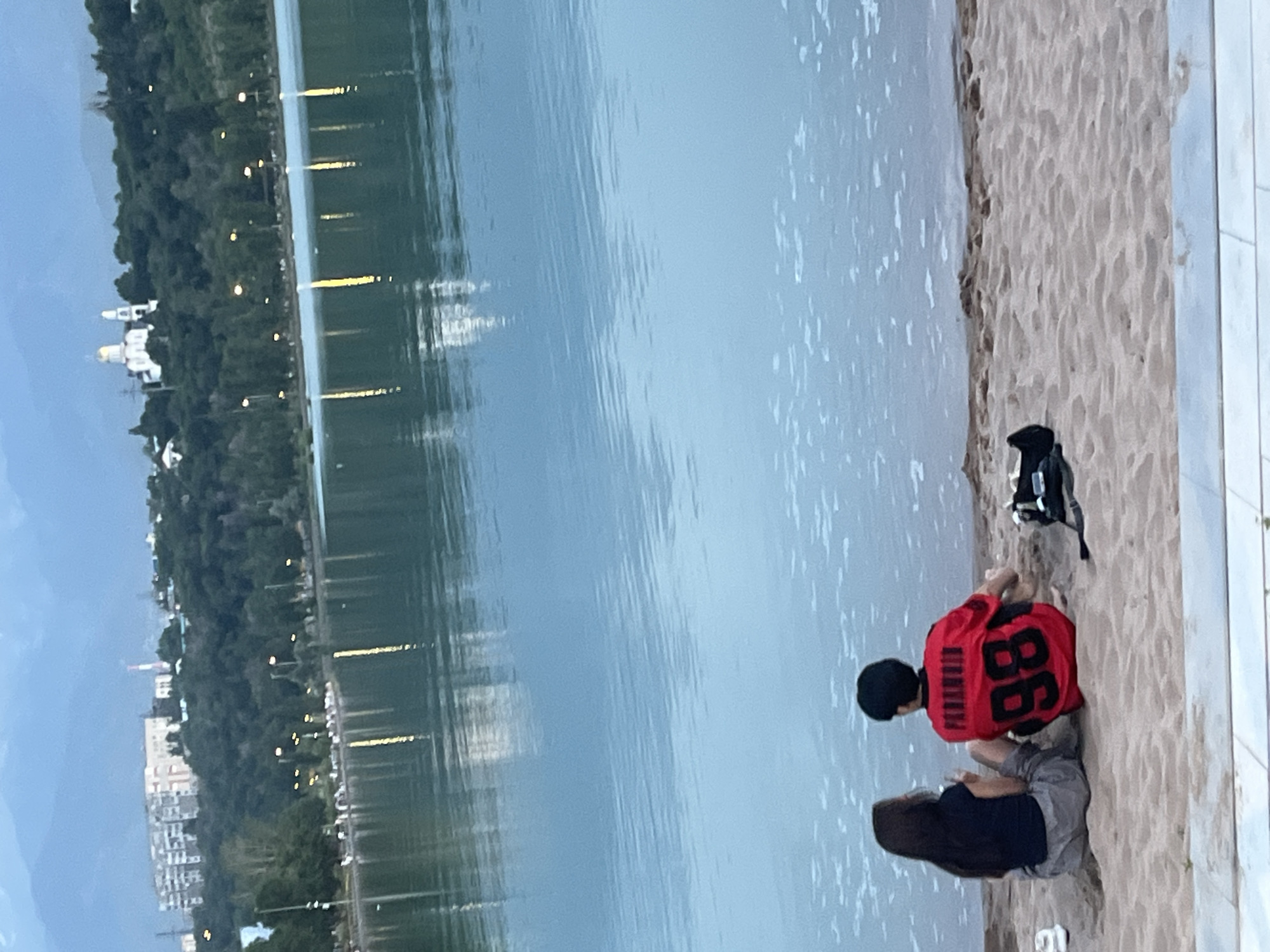 The image depicts a serene lakeside scene at dusk in Almaty, Kazakhstan. The foreground features a sandy beach where two individuals are seated near the water's edge. One person wears a red t-shirt with the number "98" prominently displayed in black. A black backpack rests behind them. The water is calm, reflecting the dim light of the setting sun and the lights along the distant shore. On the far side of the lake, a lush green treeline is visible, punctuated by scattered lights and buildings, including a distinctive white structure with a golden dome, likely a church or mosque, suggesting a cultural landmark. Further along the shoreline, apartment buildings and streetlights add to the urban backdrop. The overall mood is peaceful and tranquil, characteristic of an early evening by the water. The sky is a muted blue, indicating the transition from day to night.