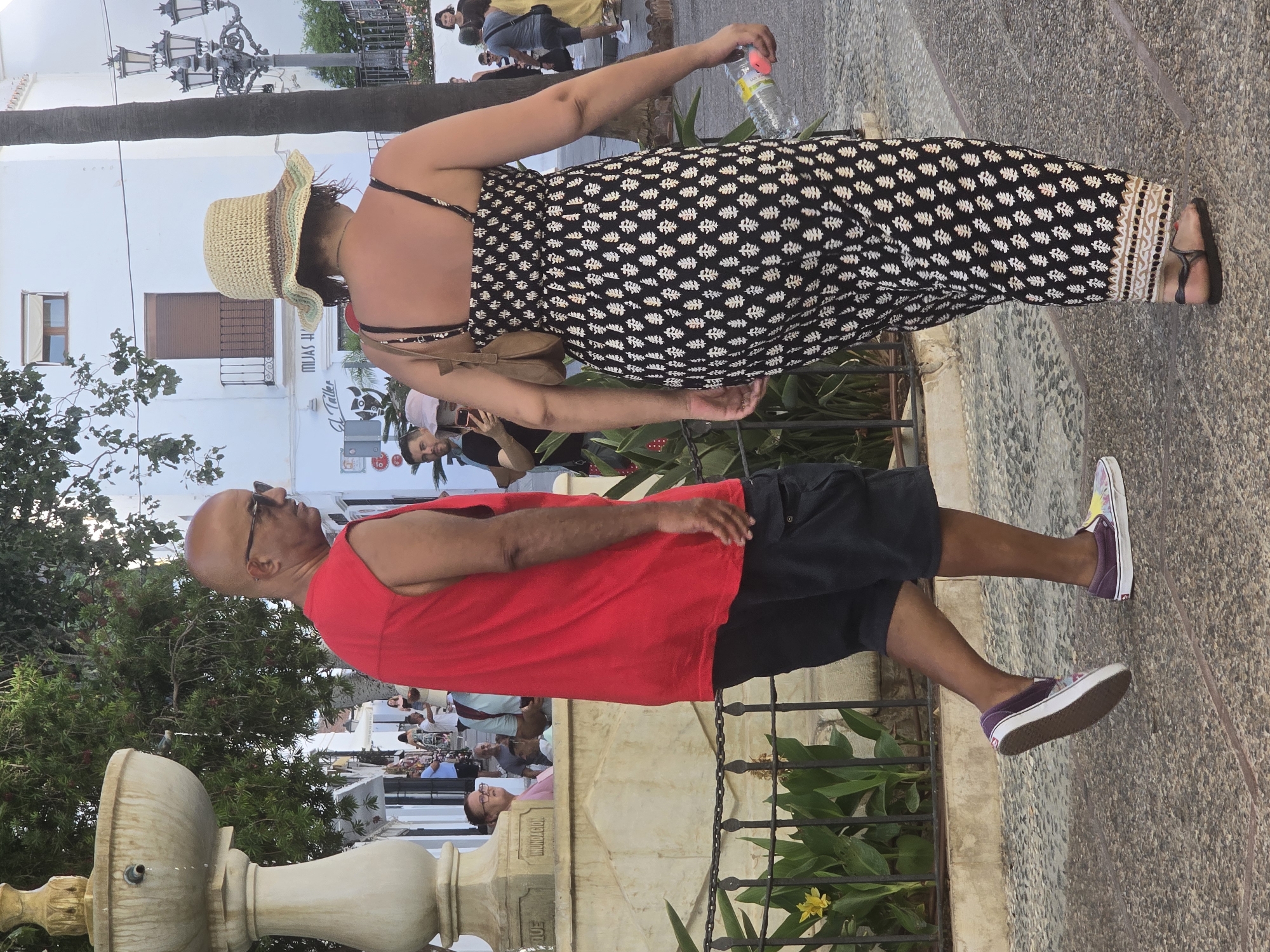 In the city of Mijas, Spain, a bald man wearing sunglasses and a red sleeveless shirt walks with a woman wearing a straw hat and a patterned dress. The man is wearing black shorts and colorful sneakers. The woman is carrying a brown purse and a water bottle. They are walking on a cobblestone street lined with palm trees and white buildings. A fountain is visible to the left of the frame. The scene appears to be a sunny day in a pedestrian area.