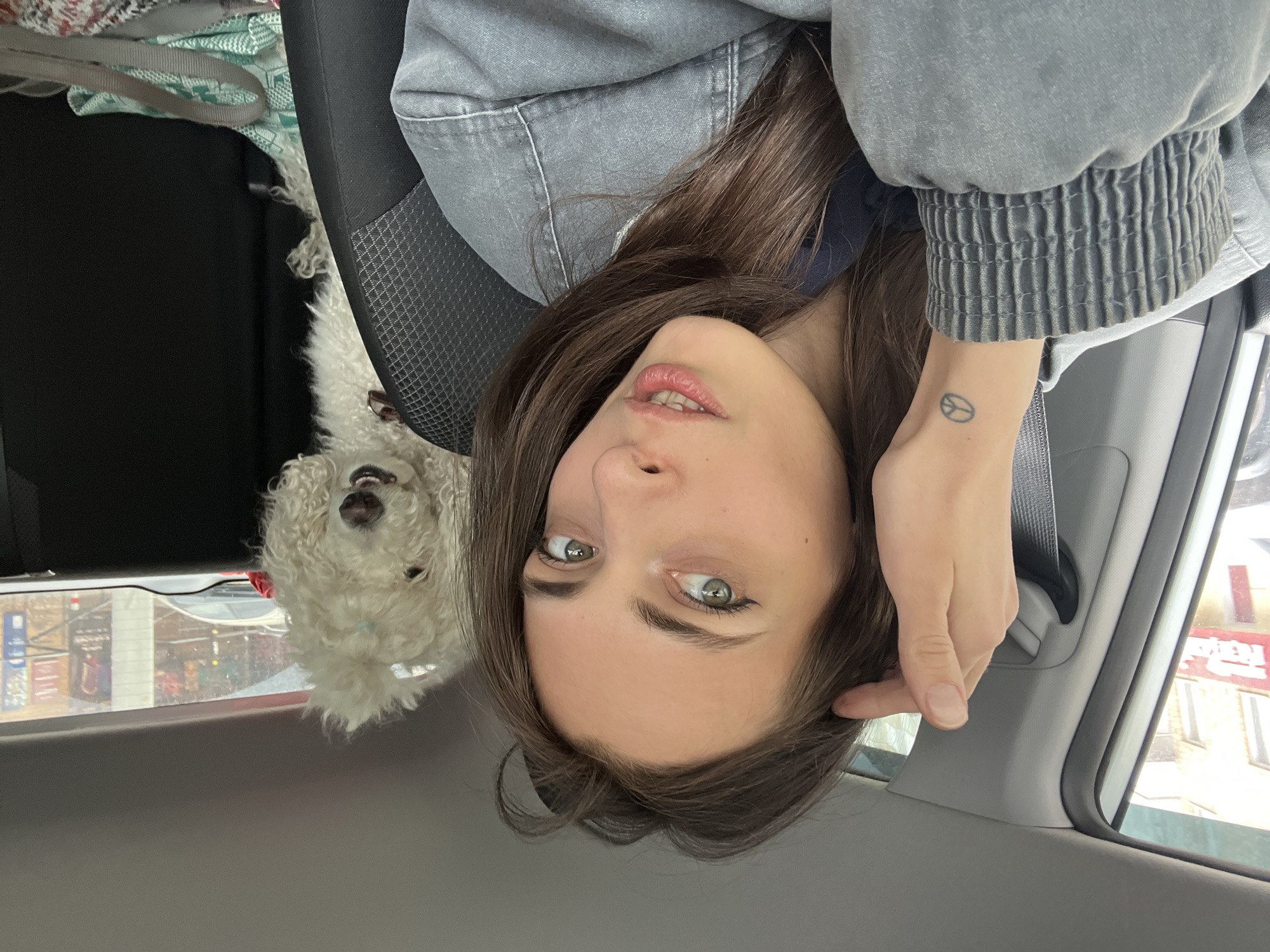 A woman with brown hair and green eyes is inside a car. She wears a grey jacket and has a peace sign tattoo on her left wrist. A white poodle is sitting next to her. Outside the car window, buildings are visible. Based on the signage in Russian, the location is in Krasnoyarsk, Russia.