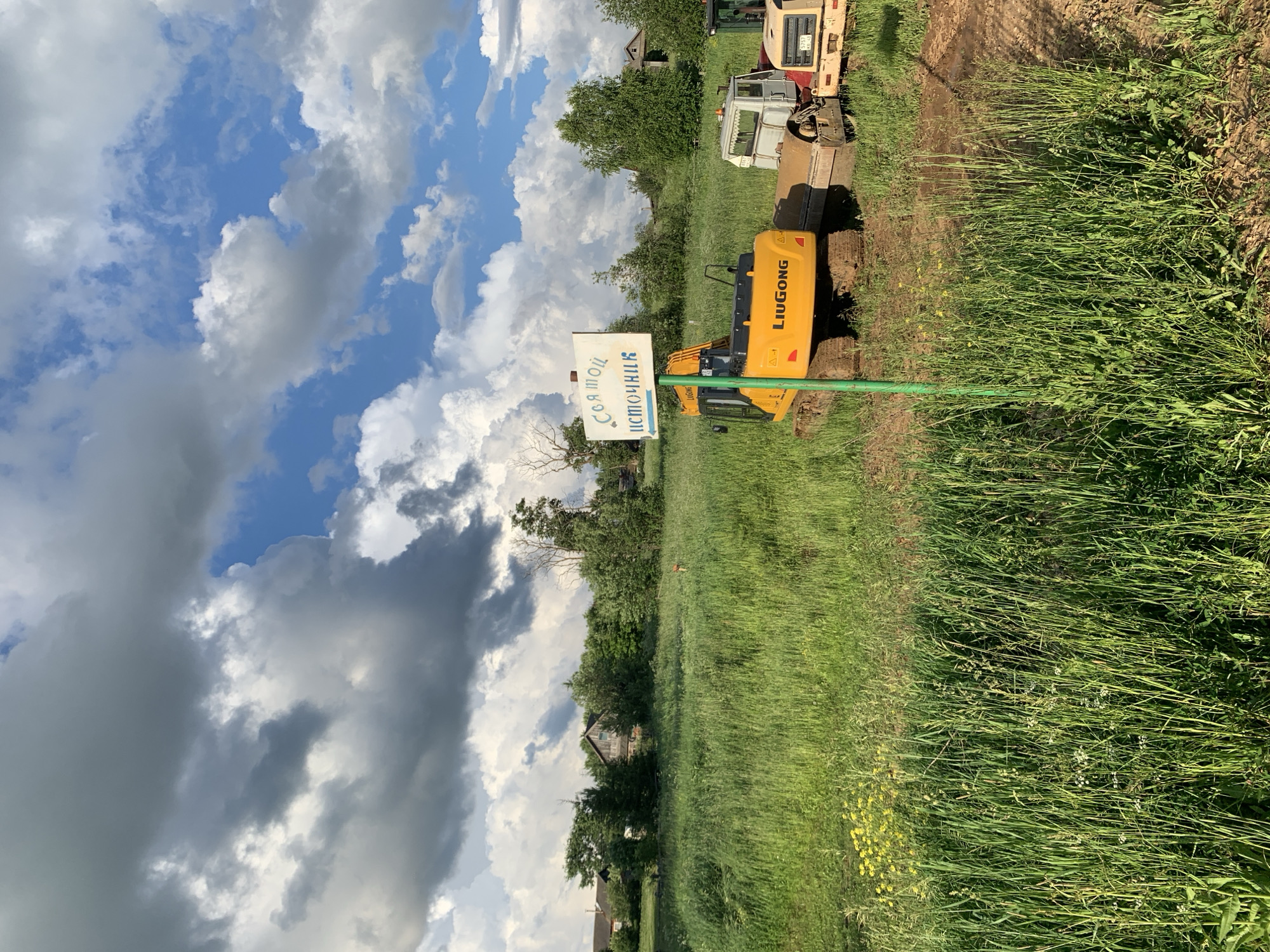 The image depicts a rural scene in the village of Svyatitsa, Russia, on a partly cloudy day. In the foreground, tall green grass dominates the landscape, with a few yellow wildflowers visible. A green signpost stands in the middle ground, bearing white lettering in Russian that reads "Соя мой источник" (Soy is my source) with an arrow pointing to the left.
Behind the signpost are several pieces of construction equipment. A yellow LiuGong excavator is positioned prominently, with its tracks resting on the muddy ground. To its right, a white truck with a raised cabin and a metal roller attachment is parked. Another piece of yellow construction equipment, possibly a bulldozer or another excavator, is partially visible to the far right.
In the background, a cluster of trees and several rustic houses suggest a residential area. The sky is filled with dramatic, dark grey clouds interspersed with patches of bright blue sky, indicating dynamic weather conditions. The overall impression is one of rural industry and construction work set against a natural, somewhat wild, backdrop. No people are visible in the frame.