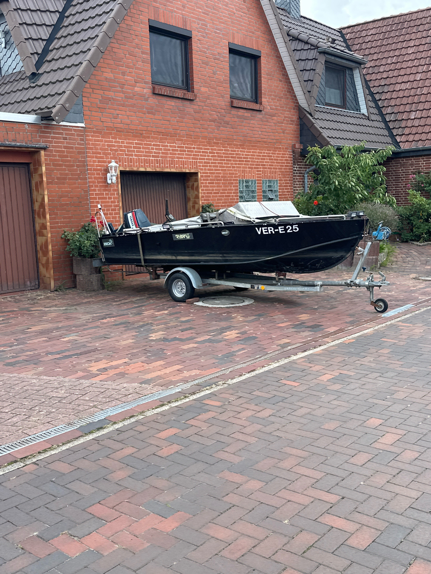 A black motorboat, identified by the registration "VER-E 25" on its stern, is parked on a red brick paved driveway in front of a brick building. The boat is mounted on a galvanized steel trailer with a single axle. A Yamaha outboard motor is attached to the back of the boat. A white cover is partially draped over the boat's cockpit. To the left of the boat, there is a tall potted plant with green foliage and a red flower. The building behind the boat is constructed of red brick with a brown garage door and a white outdoor light fixture. The roof of the building is covered with dark gray tiles. The scene appears to be a residential property in the Samtgemeinde Thedinghausen, Germany, given the presence of the boat on a trailer and its proximity to a residential building. The ground is paved with red and dark gray bricks laid in a herringbone pattern. There is a metal grate drain along the edge of the paved area. No people are visible in the image.