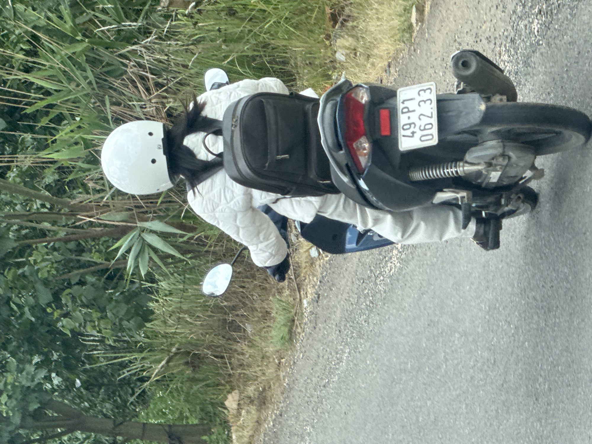 A person wearing a white helmet and a quilted white jacket is riding a dark-colored motorcycle on a paved road. They are also wearing light-colored pants and dark gloves. A black backpack is strapped to their back. The motorcycle has a license plate that reads "49-P1 062.33". The background consists of lush green vegetation, including trees and tall grasses, typical of the rural environment in Đạ Cháy, Vietnam. The road appears to be a secondary road, with a gravel shoulder. The lighting suggests it is daytime.