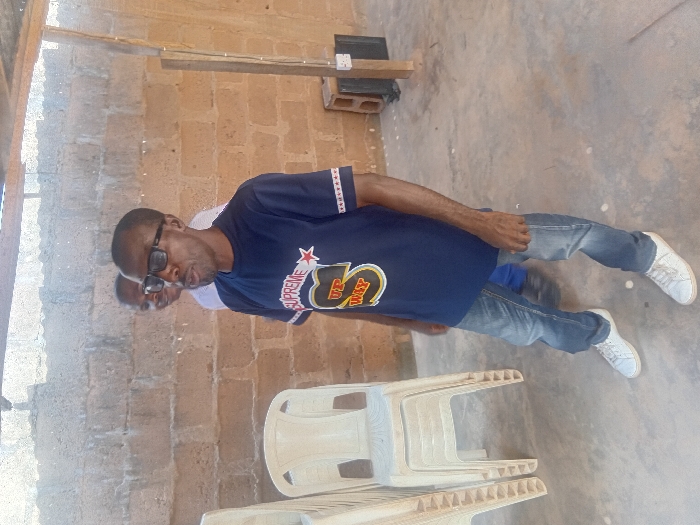 The scene depicts two adult males in a semi-enclosed, utilitarian area within Ayangba, Nigeria. In the foreground, an adult male with dark skin, short dark hair, and a slight beard, wearing black rectangular glasses, a dark blue short-sleeved t-shirt with "SUPREME IT'S MY WAY" graphics, blue jeans, and white sneakers, stands with his mouth slightly open, facing slightly to the viewer's right. His arms are down, hands loosely clasped. Behind and to the left of the primary subject, a second adult male with lighter skin tone is partially visible, appearing to smile. To the left of the foreground subject, a stack of four light-colored plastic chairs is present. The background features an unfinished light brown concrete block wall. The floor is a light gray concrete surface with scattered white debris. On the right side, a vertical wooden beam supports an overhead structure, with a black electrical outlet or box attached near its base. The area is illuminated by bright natural light.