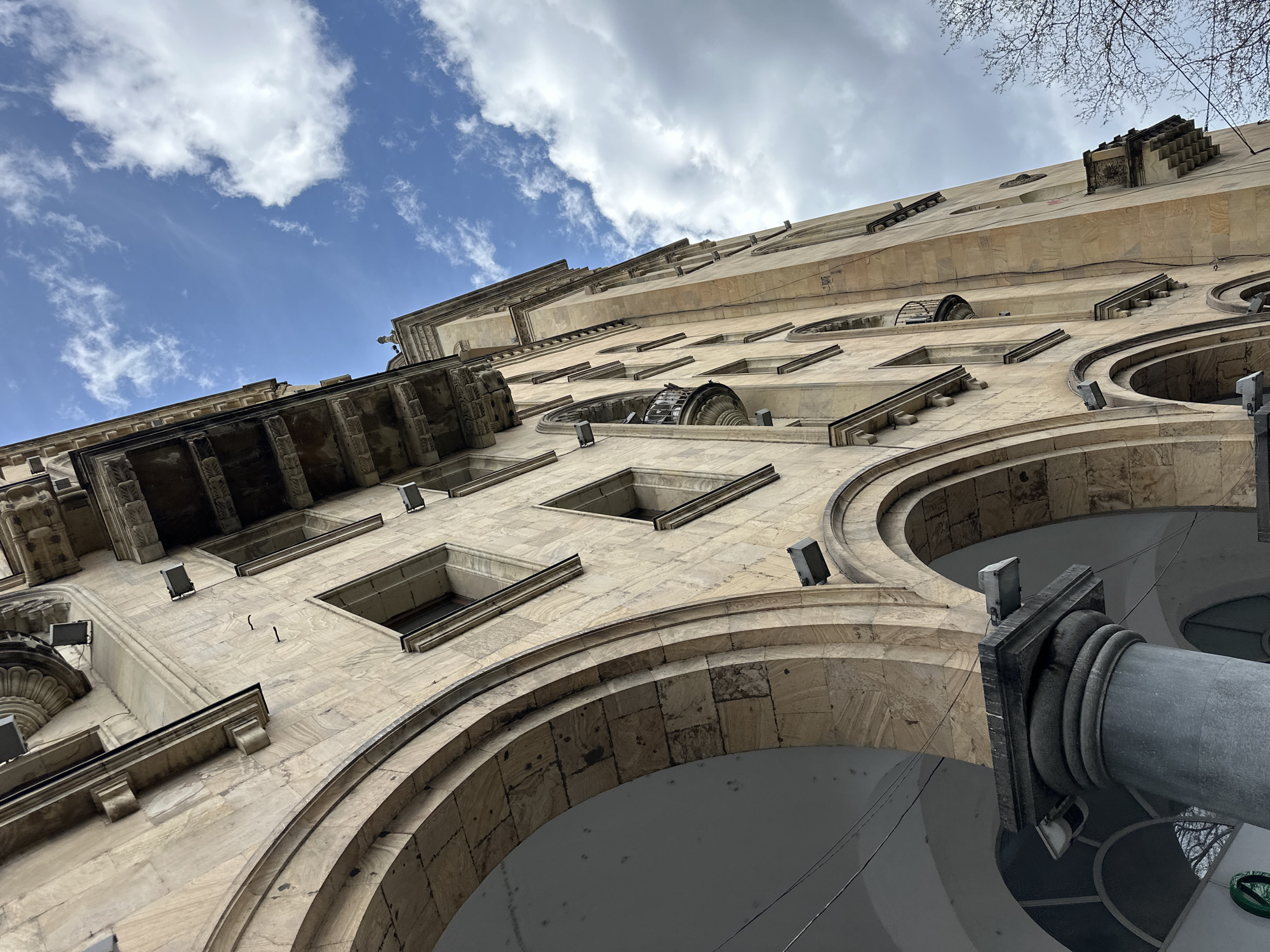 The image depicts a worm's-eye view of a historic building facade in Tbilisi, Georgia. The architecture is characterized by light-colored stone construction featuring arches, rectangular windows, and decorative embellishments. A stone column with a gray finish is prominent in the foreground, intersecting with one of the arches. The sky, visible above the building, is partly cloudy with a mix of blue and white. Bare branches from a tree are visible in the upper right corner. The building appears to be well-maintained and is lit by small, square lights affixed to the facade. The overall impression is one of classical architecture set against a vibrant sky.