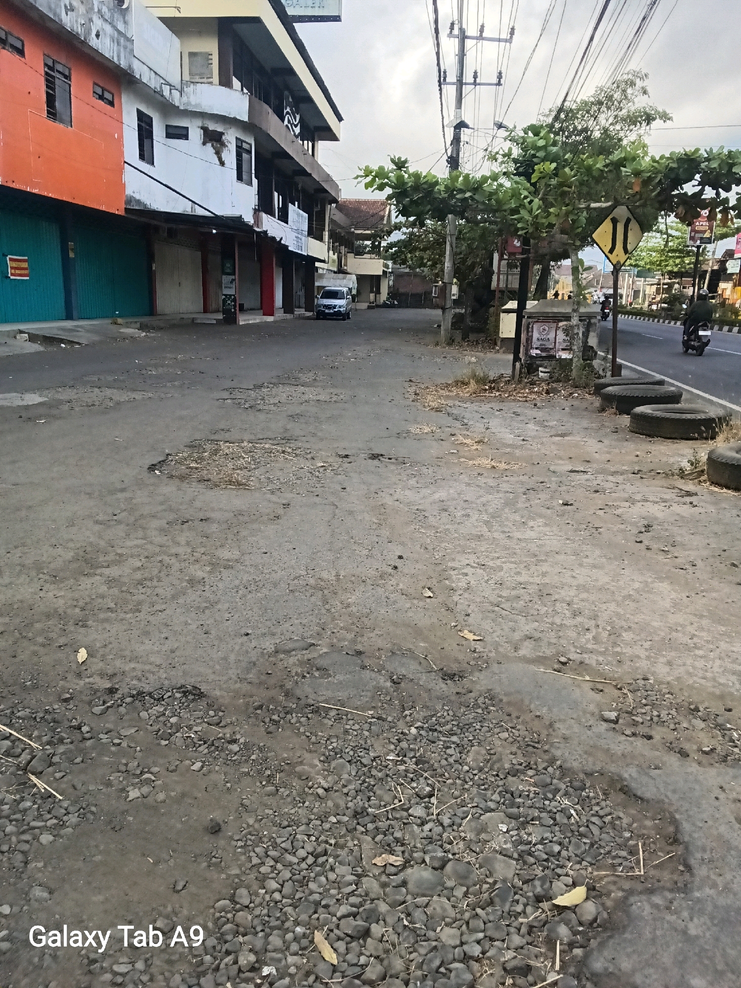 The image shows a street scene in Trenggalek, Indonesia. The foreground features a cracked and potholed asphalt road with scattered gravel and dry vegetation. In the midground, several used tires are stacked on the side of the road. A group of trees with dense green foliage stands near the road. Various buildings are visible in the background, including a multi-story structure with orange and white facades. Electrical poles and wires crisscross the sky. A silver car is parked on the side of the road. A motorcycle with a rider is seen moving along the road in the distance. A yellow diamond-shaped road sign with black markings is positioned on a pole amongst the trees. The sky is overcast. The overall scene depicts an urban street with signs of wear and tear on the road surface.