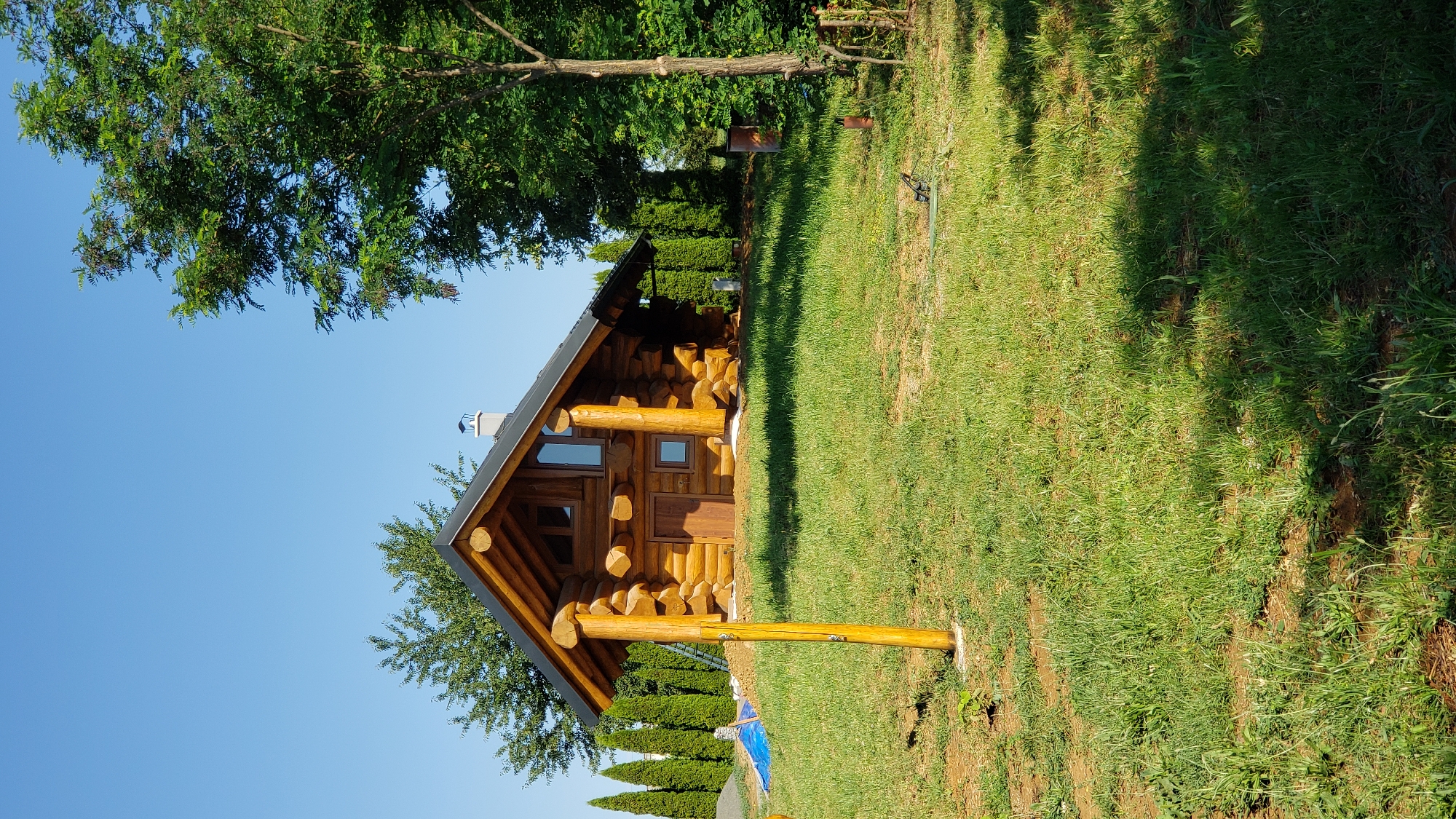 The image depicts a newly constructed log cabin situated on a grassy incline under a clear blue sky. The cabin is made of natural logs and features a dark roof with a chimney. There are windows and a wooden door on the front of the cabin. In front of the cabin, a wooden post with some attachments stands on the lawn. The surroundings are characterized by lush green grass and various trees, including tall evergreen trees lining the property. There are also some construction materials visible in the background. The overall scene suggests a tranquil, natural setting, possibly in a suburban or rural area of Prešov, Slovakia, given the architectural style and landscaping.