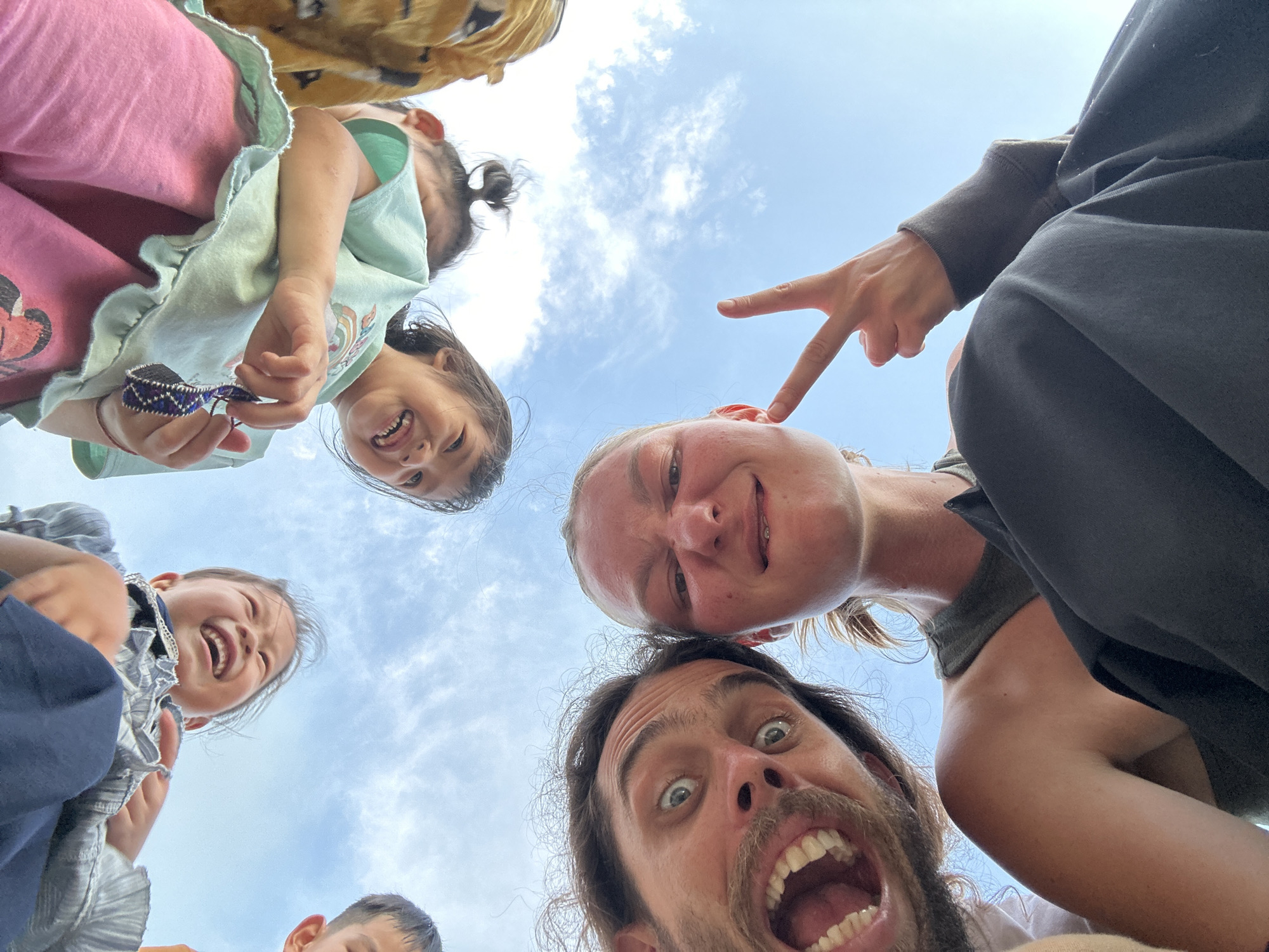 The image presents a low-angle perspective, looking upwards at a group of six individuals against a backdrop of a clear blue sky with scattered white cumulus clouds.
In the immediate foreground, two adults occupy the lower half of the frame. On the left, a man with a dark beard and mustache is captured with his mouth wide open, revealing teeth, and his eyes wide, conveying an expression of exultation or surprise. He wears a light-colored, possibly white, garment. To his right, a woman with light-colored hair pulled back makes a "peace" sign with her left hand while her face is contorted into a playful, scrunched expression. She wears a dark olive green sleeveless top and dark-colored pants.
Above and behind the adults, four children are partially visible, oriented as if looking down towards the camera. One child at the top-center wears a grey and blue ruffled top and displays a wide, open-mouthed smile. To the upper-right, another child with dark hair tied in a bun wears a light green ruffled top, possibly with a colorful emblem, and holds a small, purple, woven or beaded object in their hand, smiling. Portions of two other children's faces are visible on the far left and far right edges of the upper frame.
The overall scene conveys a playful and joyful interaction. The specific location within the city of Gia Hội, Vietnam, is not visually discernible from the image content, which shows only the sky.