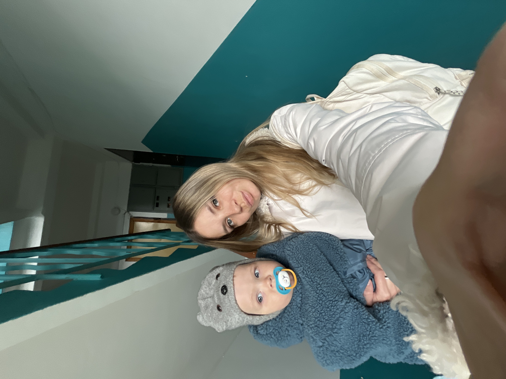The image displays a woman and an infant, captured from a high-angle perspective looking downwards, with the photo presented rotated 90 degrees clockwise. This intimate indoor scene takes place in Chaikovsky, Russia.
In the upper portion of the frame (as rotated), a woman with long, straight blonde hair looks directly at the camera with a neutral expression. She is wearing a white, possibly padded, jacket or coat. Her body is positioned diagonally across the frame, suggesting she is holding or leaning over the baby.
The baby, positioned below the woman, is wearing a distinctive grey knit hat with bear-like ears and a blue, textured fuzzy jacket. A light blue pacifier with a yellow/orange ring is in the infant's mouth, and the baby's bright blue eyes are wide open, looking towards the camera. The woman's hand is partially visible, seemingly supporting the baby.
The background reveals an interior space, characterized by walls painted in two distinct sections: a lighter, possibly off-white or light grey, upper portion and a vibrant teal or turquoise lower section. A teal-colored railing, matching the wall color, runs diagonally across the lower left area, suggesting a stairwell or landing. The lighting indicates an indoor setting, possibly with artificial illumination. There is no visible text in the image.
