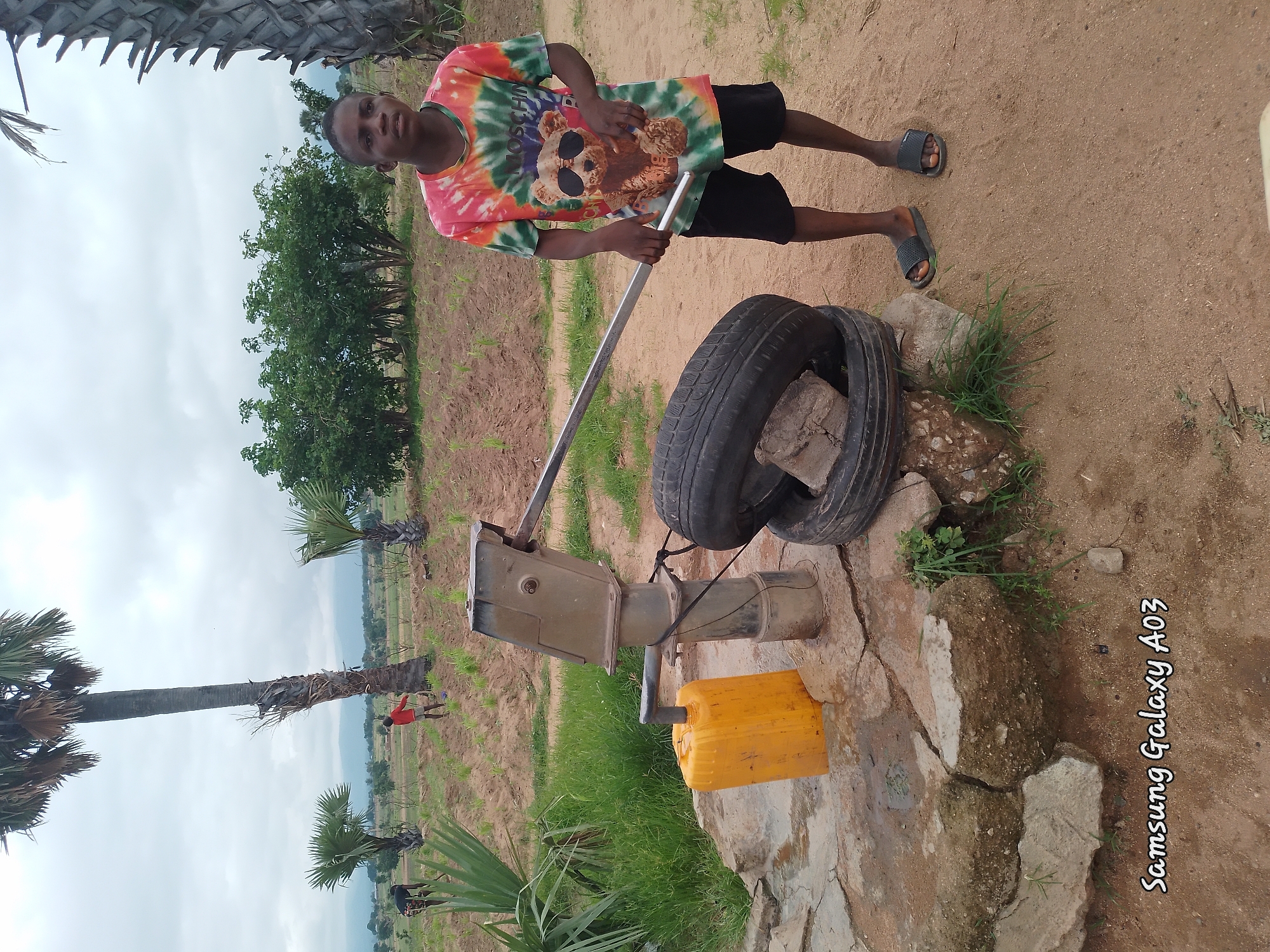 In Angwan Hausawa, Nigeria, a young boy stands by a water pump. The boy wears a tie-dye t-shirt with a graphic of a teddy bear in sunglasses and black shorts, along with dark flip-flops. In front of him, two car tires are stacked, with a piece of concrete resting on top of them. To the left of the tires is a yellow plastic water container connected to the pump's spout. The water pump itself is metallic and mounted on a stone base. The ground is sandy and uneven, with scattered rocks and patches of green grass. In the background, a field with young plants stretches towards a line of trees and palm trees under a cloudy sky. Several other figures, possibly children, are visible working or playing in the distance. The overall environment suggests a rural or semi-rural setting, possibly a village or farm.