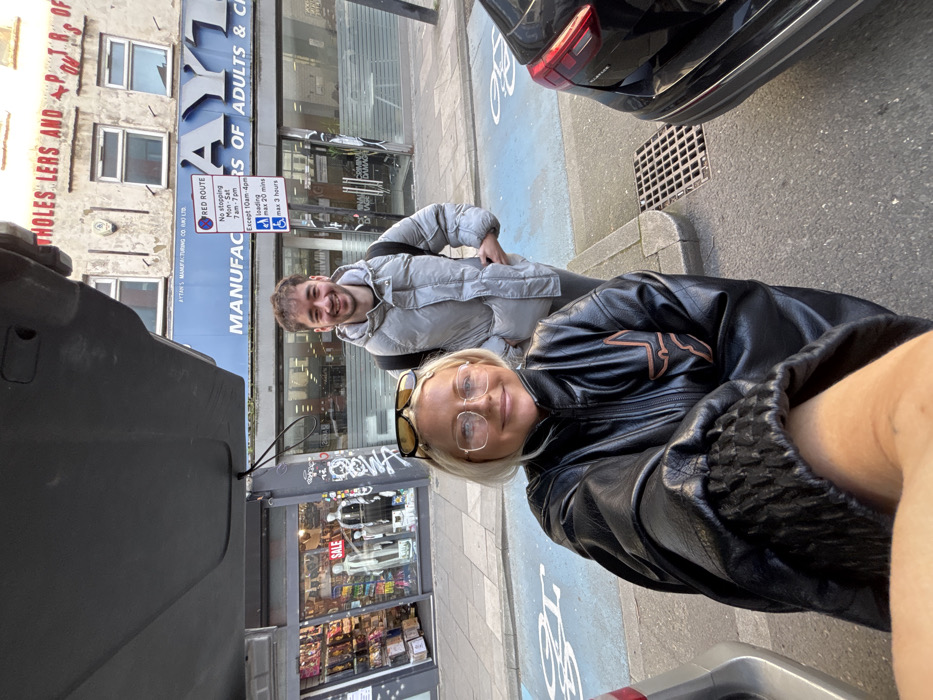 An image depicts two individuals, a female and a male, posing for a selfie on a street in London, United Kingdom.
The female in the foreground, with blonde hair and clear-framed glasses, smiles directly at the camera. Yellow-tinted sunglasses are pushed onto her head. She wears a black zip-up jacket, possibly faux leather, with a distinct brown/orange stylized logo on the left chest and a textured cuff on her extended right arm, holding the camera.
Behind her, to the right, a male with short dark hair and stubble smiles at the camera, hands in his pockets. He is wearing a light grey puffer jacket, dark trousers, and a dark backpack.
The scene is set on a pavement adjacent to a road. Visible street markings include blue painted sections, likely indicating a cycle lane or parking, and a metal drain cover. The perspective of the photograph appears to be from the interior of an open vehicle boot or hatchback, with a dark underside structure and a cable visible in the top left.
In the background, multiple commercial establishments line the street. Directly behind the individuals is "AYTAN'S MANUFACTURERS OF ADULTS & CHILDREN'S...", identifiable by a large blue sign. Above this, a worn building facade shows "WHOLESALERS AND SUPPLIERS OF..." in faded red text. A prominent red "RED ROUTE" sign is visible, displaying "No stopping Mon-Sat 7am-7pm, Except 10am-4pm. Loading max 20 mins, max 3 hours." to its right. To the left of Aytan's, another shop window displays mannequins and a "SALE" sign. Black graffiti is present on a pillar between the shops. A black car is partially visible on the right, parked parallel to the kerb.
