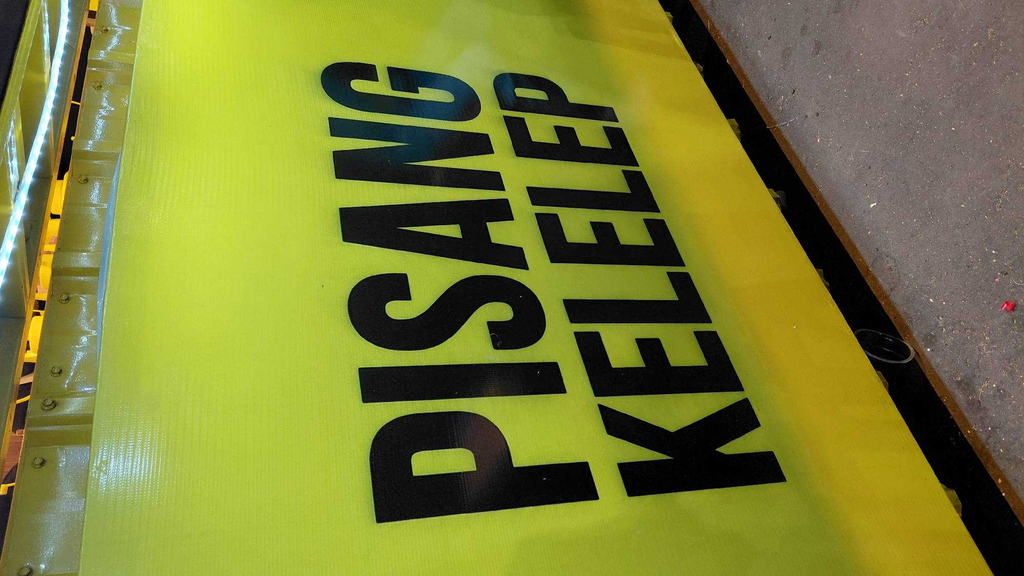 A bright yellow, corrugated plastic sign with bold black lettering reading "PISANG KELELEP" is mounted on the side of a structure. The sign is positioned at eye level and takes up most of the frame. Above the sign, a yellow metal structure is visible with several bolts and a strip of white LED lights running horizontally. The ground below the sign is a mix of dark material and textured concrete or asphalt, with some debris scattered on it, including a small piece of red plastic. The lighting suggests it is nighttime or a dimly lit area. The overall impression is that of a food stall or vendor. The sign likely refers to a type of banana fritter or dessert. The location is specified as Bantarsoka, Indonesia.