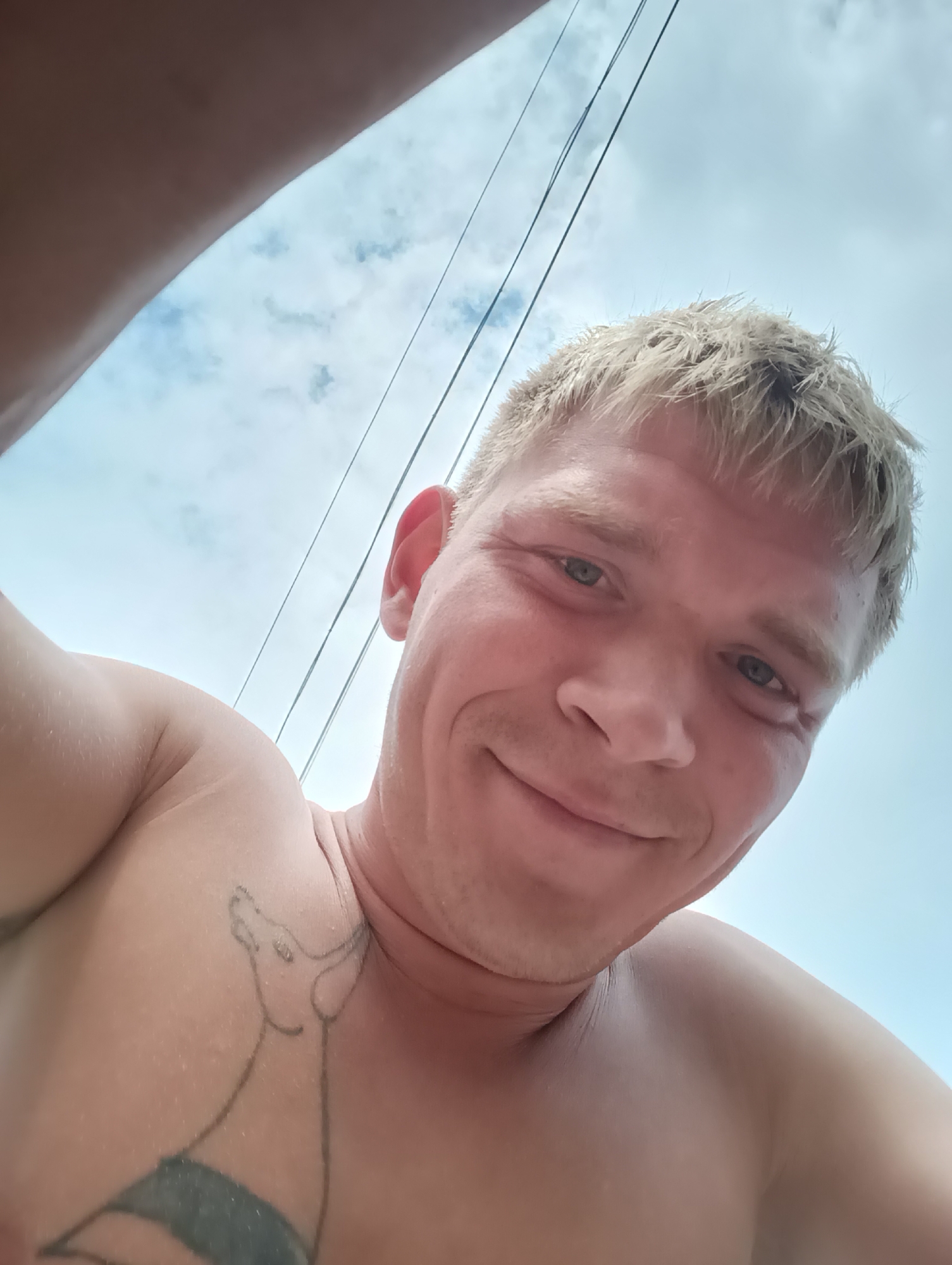 A close-up, low-angle, selfie-style photo shows a shirtless Caucasian male with short, spiky, blonde hair. He is smiling and looking directly at the camera. His head is tilted slightly upwards, and his left ear is visible. A tattoo of a stylized animal, possibly a dog or a horse, is visible on his upper left chest. The background consists of a bright blue sky with scattered clouds, and several thin dark wires are visible, suggesting an outdoor environment possibly with utility poles or overhead wires. The lower left portion of the image is obscured by a dark, fleshy shape, likely the arm or hand of the person taking the photo. There is no identifiable information in the image to suggest a specific location within the city of Chaikovsky, Russia.