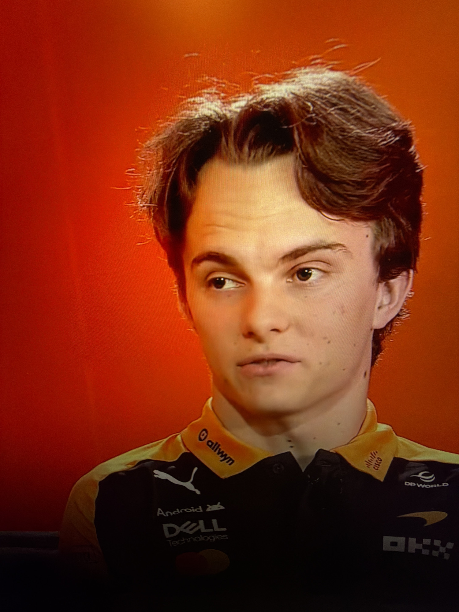 The image is a close-up, medium shot of a young man with brown hair against an orange background. He is wearing a black collared shirt with yellow accents and several logos. On the collar, there are logos that appear to read "allwyn" and "CISCO". Below the collar, there are logos for "Puma," "Android," "Dell Technologies," and "DP WORLD," as well as a stylized arrow logo. The man is looking slightly off to the left of the frame, with his mouth slightly open. His facial features include light skin, brown eyes, and a few freckles. His hair is styled with some loose strands. The overall lighting suggests an indoor setting, possibly a studio or a television broadcast. There is no visual information in the image that indicates the city of Kafr Yasif, Israel.