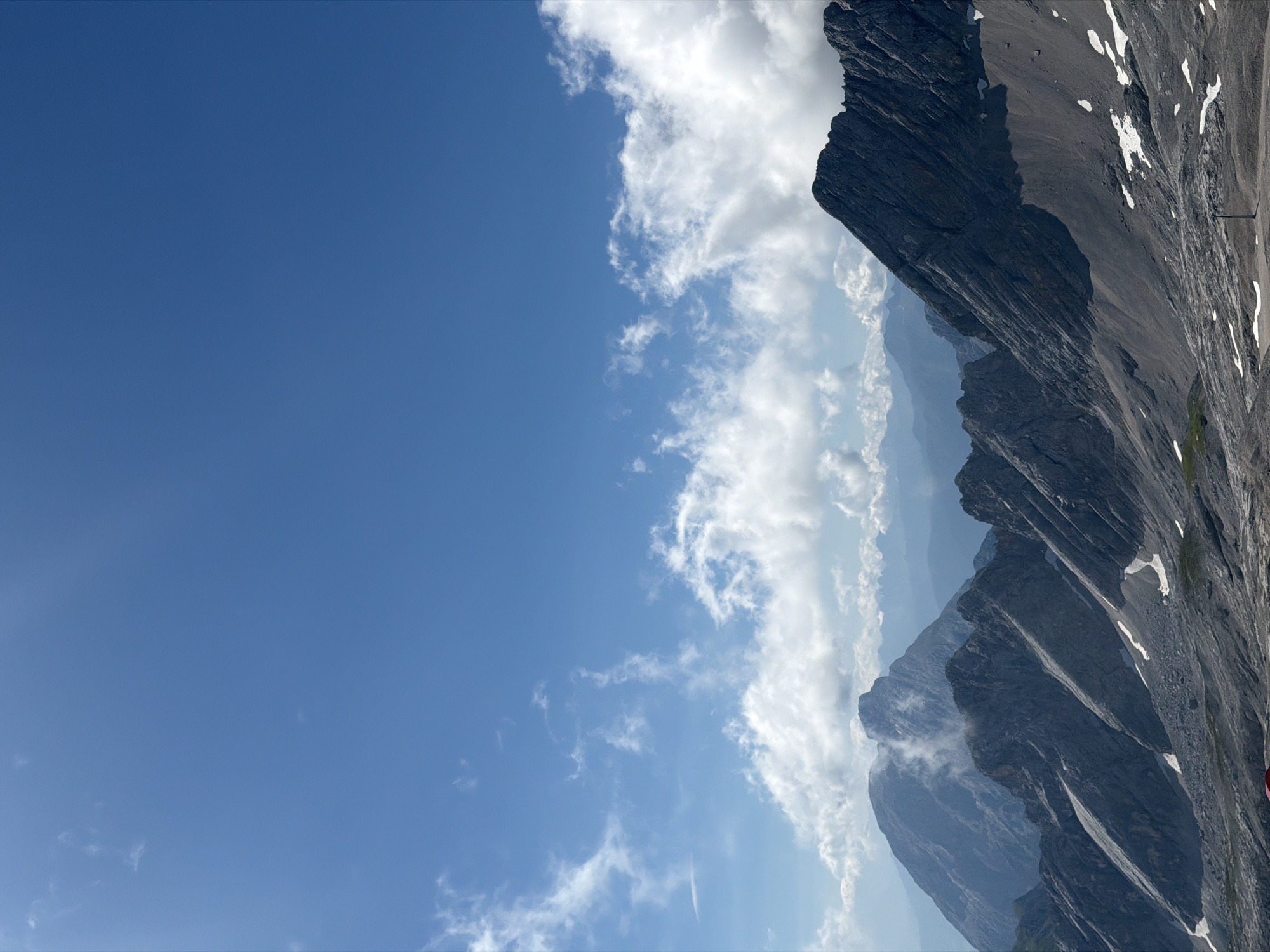 This is a high-angle, medium-shot, outdoor, vertical, natural landscape photograph taken on a clear, sunny day. The scene depicts a vast mountain range under a bright blue sky dotted with scattered white clouds. The foreground is dominated by a rugged, rocky mountain slope with patches of snow. In the middle ground, more sharp, angular mountain peaks rise, some partially obscured by mist. Further in the background, a series of softer, blue-hued mountain ranges recede into the distance. A small, red tent is visible in the lower left corner of the image. The overall impression is one of a remote, alpine environment. The photograph is likely taken from a high vantage point, possibly a mountaintop or a viewpoint. While Wildermieming, Austria, is mentioned as a location, there are no specific landmarks or urban elements within the image to confirm this with certainty.