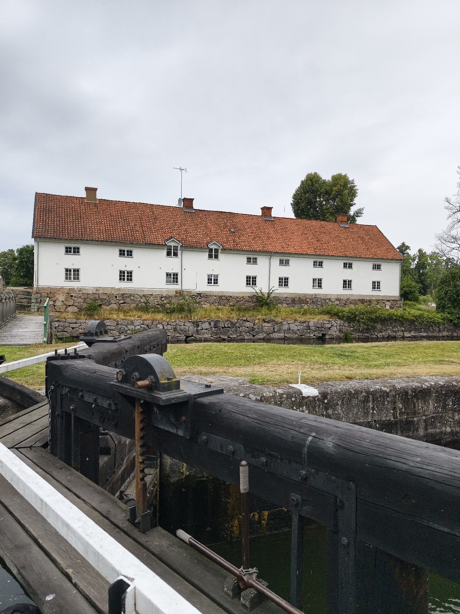 This image depicts a lock gate in the Göta Canal at Brokind, Sweden. The lock gate, constructed from dark wood, is in the foreground, with a water channel visible below it. Behind the gate, a stone wall borders a grassy area. Further back, a long, white building with a red-tiled roof stands on an elevated embankment. The building features multiple windows and chimneys, and its facade is punctuated by dormer windows on the upper story. Trees line the background, with an overcast sky overhead. The scene appears to be serene and devoid of human activity.