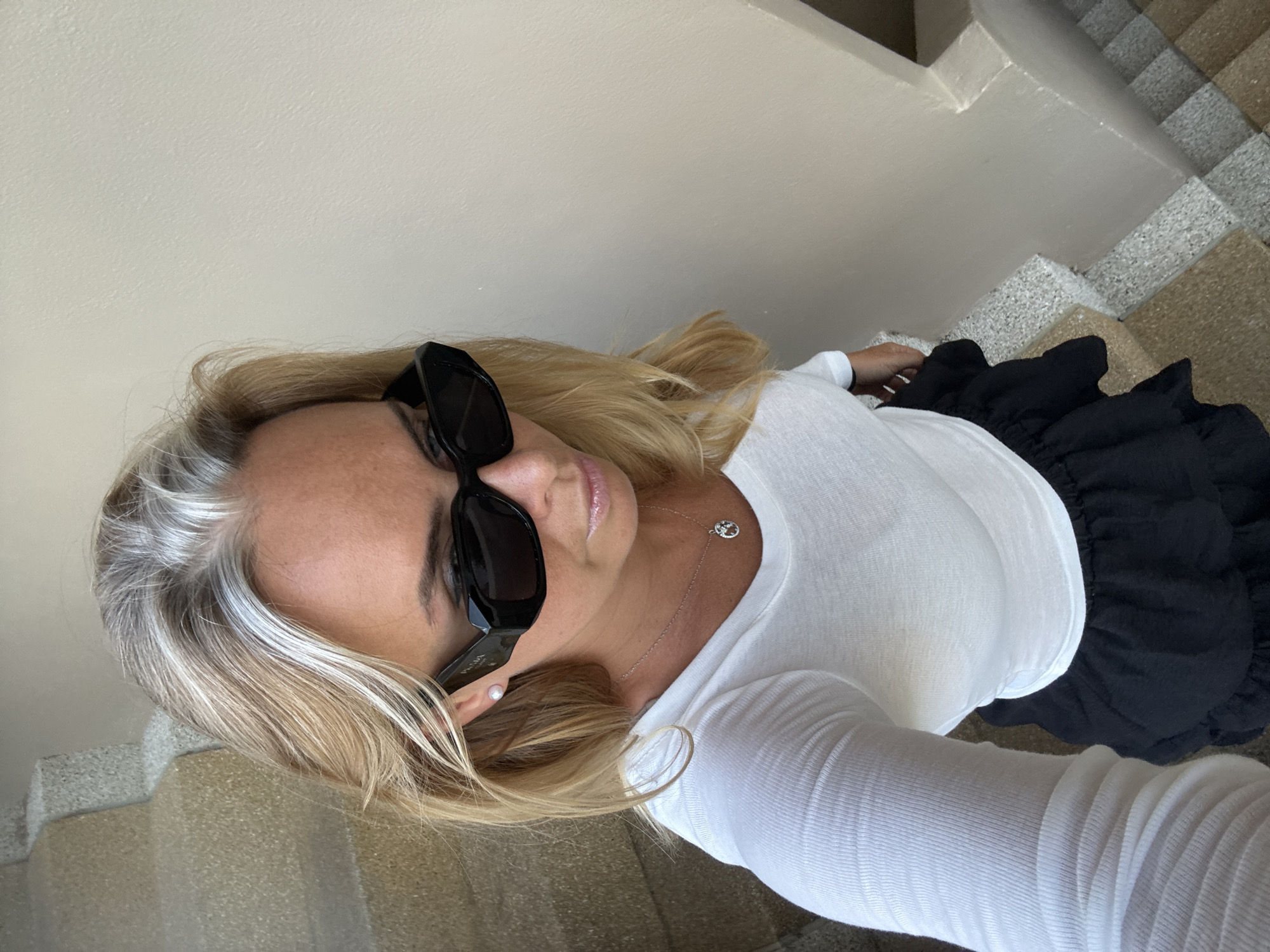 A woman stands on a staircase in a building, possibly a hotel or residential complex, in Baan Chaweng, Thailand. She is wearing a white long-sleeved top, a black ruffled skirt, and black sunglasses. The staircase has speckled grey and yellow steps and a cream-colored wall to her right. She is holding a phone in her left hand, presumably taking a selfie. Her blonde hair has streaks of grey and is styled loosely around her shoulders. She is also wearing a pearl earring and a silver necklace with a pendant. The lighting suggests it is daytime.