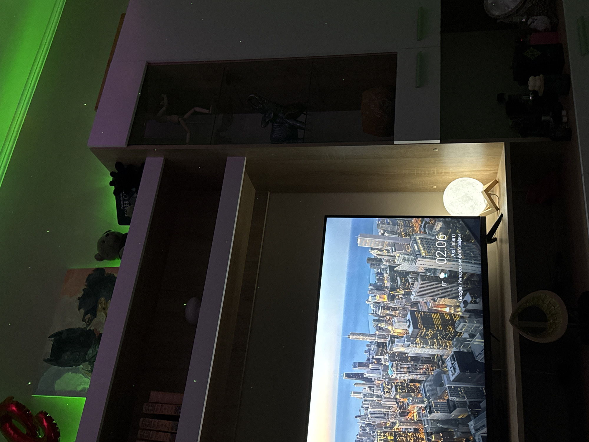 This dimly lit interior scene showcases a modern entertainment or shelving unit with various decorative and functional items, illuminated by ambient lighting. The perspective is tilted, with the main subject being a large flat-screen television displaying a vibrant cityscape.
On the left side of the room, green LED strip lighting casts a glow along a wall or ceiling edge. Below, a red foil balloon, possibly shaped into letters, is partially visible. A framed artwork or poster depicting a dark-clad figure, resembling Batman, is displayed on a shelf, above which a small teddy bear rests. Further down, a black "Proboxlab POKER C+" device is placed on a shelf, and several stacked books are visible at the bottom. A glass-fronted cabinet above holds a wooden articulated mannequin in a dynamic pose, a dark, possibly reptilian sculpture, and a spherical orange object.
On the right side, above the television, a moon-shaped LED lamp on a wooden stand emits a soft glow. The television screen prominently displays a detailed cityscape as its wallpaper or screensaver. Visible text on the screen indicates the time "02:06", a temperature of "8°", the name "Asif Islam", and "Google • Интересные фотографии" (Google • Interesting photos), suggesting the source of the background image. To the immediate right of the television, a light-colored, possibly woven, hat or bowl-like object is present. The top shelves of the unit contain various dark, indistinct objects, including what appears to be a woven basket. The overall setting is a domestic space, likely a living room or bedroom, captured in the early morning hours, with no specific activity or individuals present.