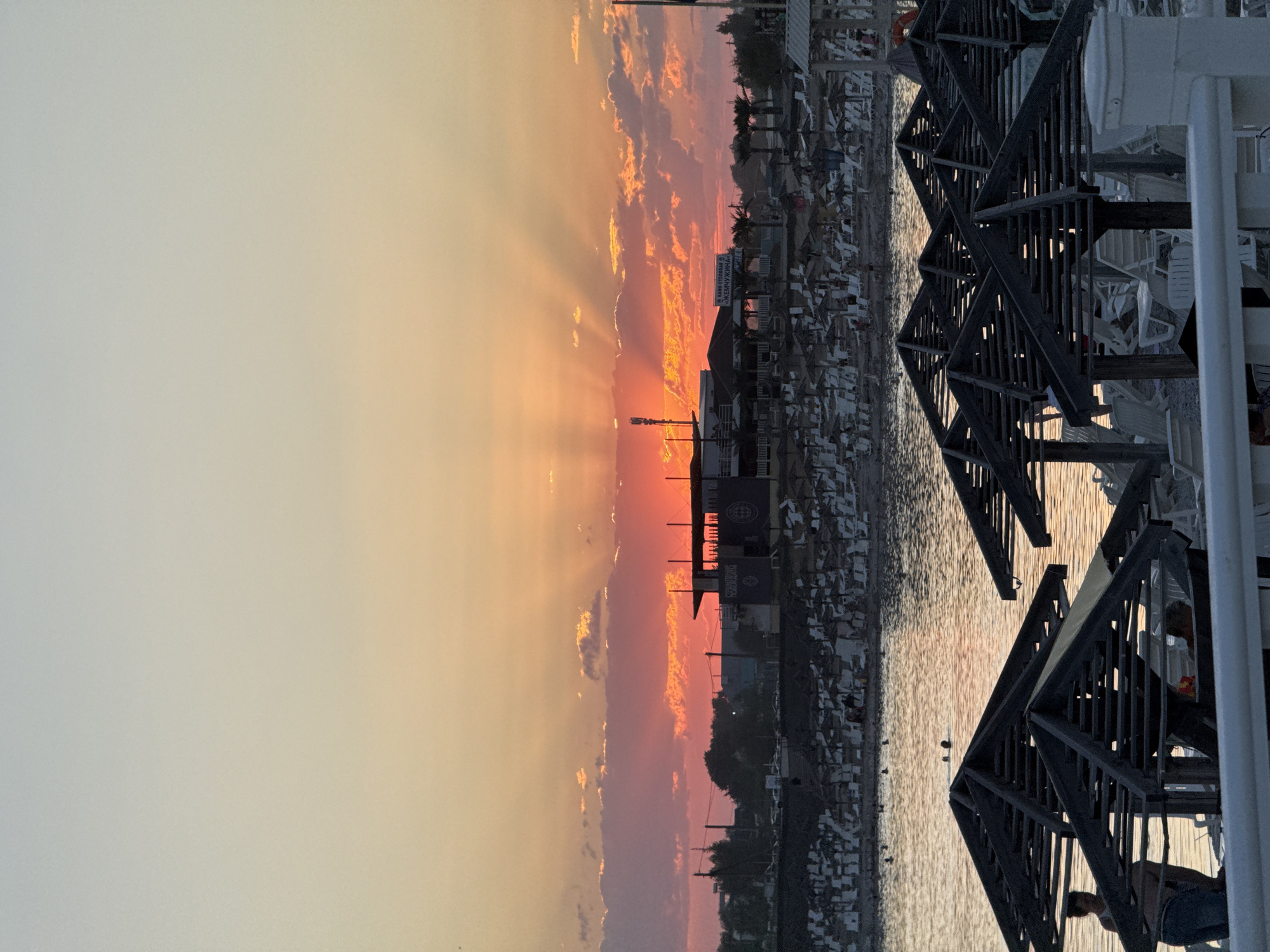The image depicts a sunset scene at a beach, likely in Sol-Iletsk, Russia, given the presence of Cyrillic signage. The sky transitions from a pale blue at the top to vibrant shades of orange, pink, and red near the horizon, with sunbeams fanning out from behind a cloud bank. In the mid-ground, a building complex with a covered outdoor seating area and what appears to be a restaurant or cafe is visible, with signage in Cyrillic. In front of this building, rows of empty beach chairs and umbrellas are set up on the sand. The foreground is dominated by dark, angular wooden structures that appear to be covered seating or shade areas for beachgoers, with several white lounge chairs underneath. In the water, a few people are visible, some swimming or wading. The sea is calm, reflecting the warm colors of the sunset. The overall impression is one of a tranquil evening at a seaside resort.