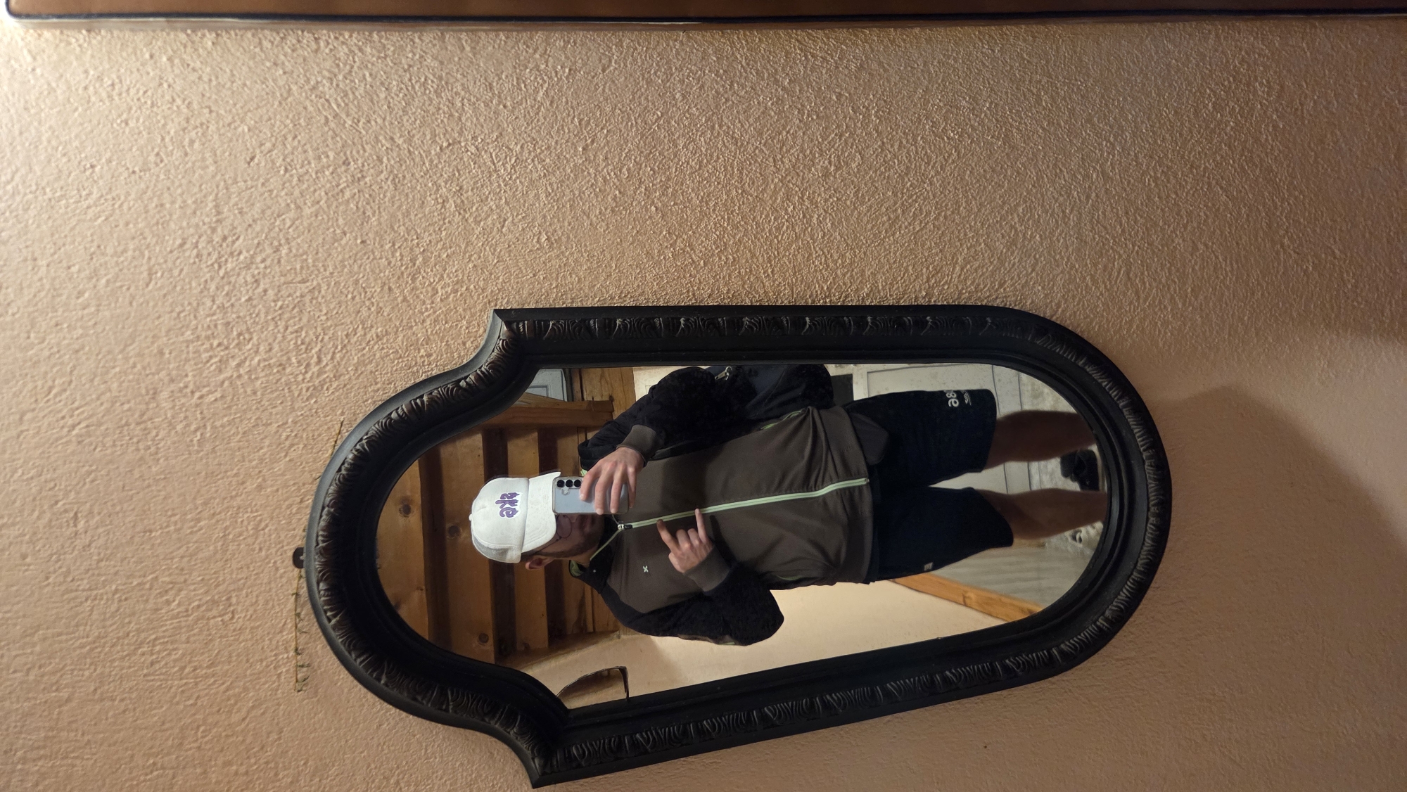 A man is taking a mirror selfie in a room with textured peach-colored walls. He is wearing a white baseball cap with purple lettering, a brown zip-up jacket with black patterned sleeves, and black shorts. He is holding a smartphone in his right hand, angled to capture his reflection. His left hand is raised, with his index finger and pinky finger extended, forming a rock and roll hand gesture. In the background, visible in the mirror, is a wooden ceiling and part of a wall. The mirror itself has an ornate black frame.