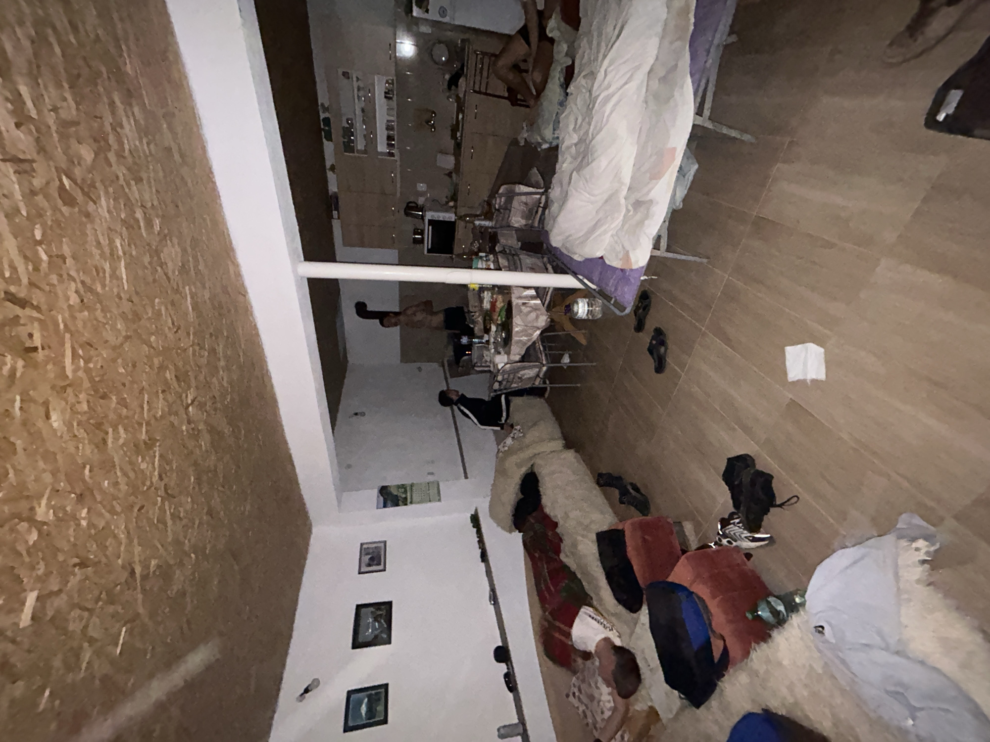 The image shows an interior scene in what appears to be a residential building, possibly in Zlatograd, Bulgaria, as requested. The room features a combination of living and dining areas, with a kitchen visible in the background. There are at least two individuals present: one appears to be standing near a dining table laden with objects, and another seems to be partially obscured, possibly lying down or resting near a couch area. The dining table has various items on it, including bottles, and possibly food. The flooring consists of wooden planks. There are personal items scattered around, including clothing and shoes. The walls are a mix of white and what seems to be unfinished wood panelling, and there are framed pictures on one wall. The room appears cluttered and lived-in.