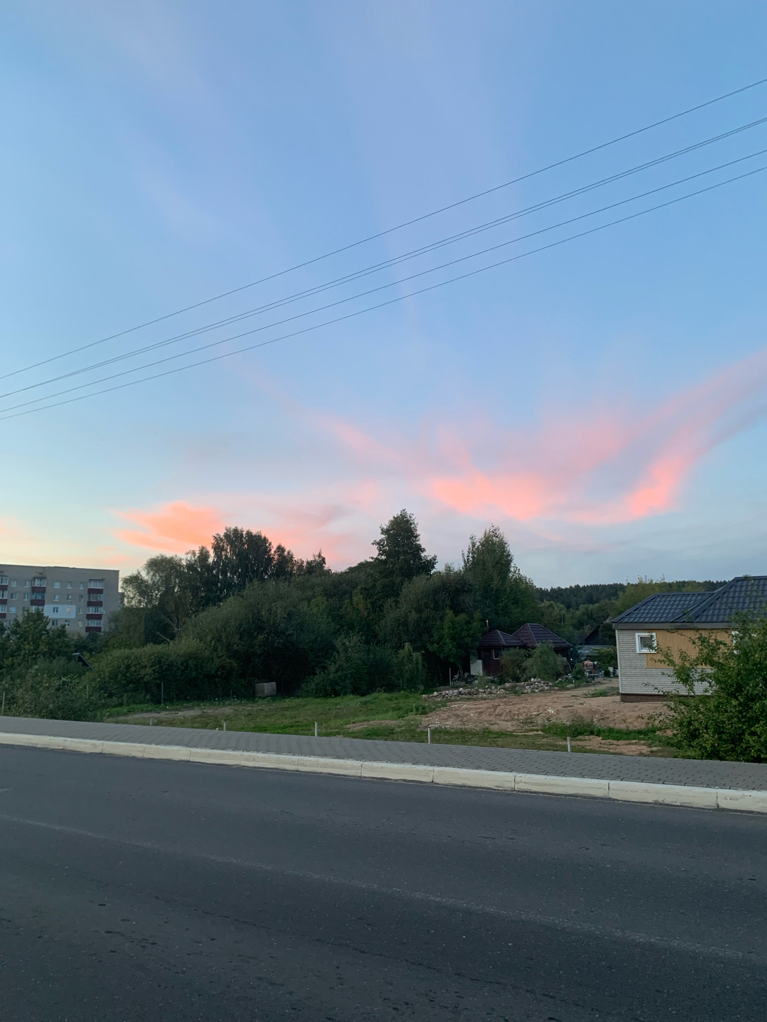 The image captures a serene sunset scene in Kapyĺ, Belarus. In the foreground, a paved road runs horizontally, with a sidewalk and curb separating it from a grassy area. Beyond the road, a line of dense green trees forms a natural barrier, with a few scattered houses and a multi-story apartment building visible to the left. The sky above is a canvas of soft blues and pinks, painted with wispy clouds that catch the warm hues of the setting sun. Power lines stretch across the sky, adding a touch of urban infrastructure to the otherwise natural landscape. The overall atmosphere is tranquil and picturesque.