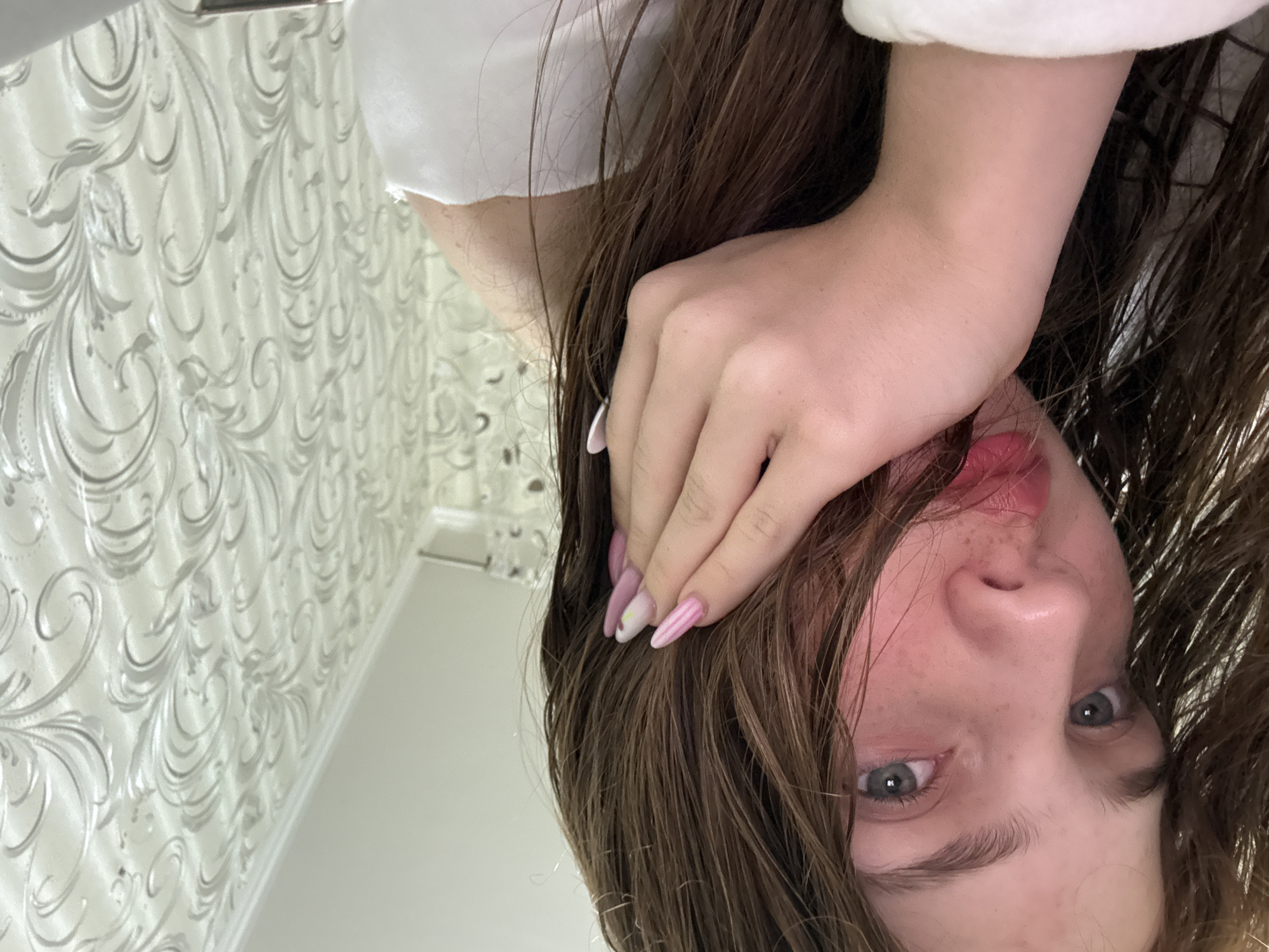This image depicts a young female, situated indoors in what is presented as a location within the city of Naberezhnye Chelny, Russia. The subject is shown from the chest up, with her head slightly tilted and her gaze directed towards the viewer. Her long, brown hair appears wet and somewhat disheveled, with strands falling across her face. Her skin is fair, exhibiting a natural flush of redness on her cheeks and nose, where small freckles are also visible. She has blue-grey eyes and full lips with pink-red lipstick. Her right hand, featuring nails painted in pink and white with subtle patterns, rests against the side of her face, intertwined with her wet hair. She is wearing a white garment, possibly a towel or a soft top, visible over her shoulder and arm. The background consists of a wall covered in textured wallpaper featuring an ornate, silvery-grey scroll and floral pattern against a light, possibly off-white, background. A white ceiling with a decorative molding strip is visible at the top right of the frame. The scene is evenly lit, indicating an indoor environment.