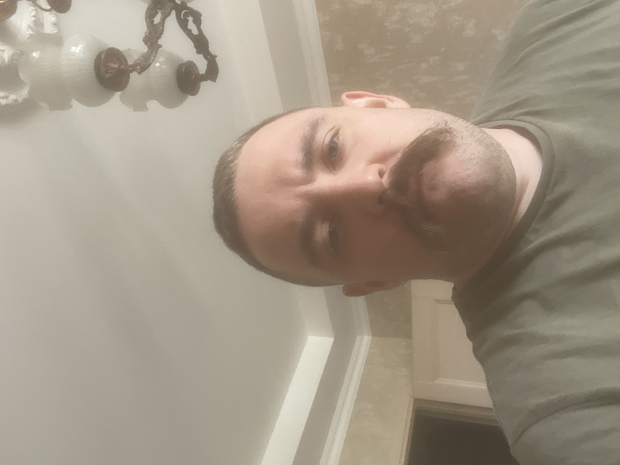 A close-up, waist-up selfie is captured from a low angle. The subject is a fair-skinned male with a buzzed haircut and a prominent handlebar mustache. He is wearing a plain olive-green t-shirt. The background features a textured, beige wall with ornate crown molding and a white ceiling. To the left, a doorway is partially visible. A chandelier with white globes hangs from the ceiling on the right side of the frame. The overall lighting is soft and diffused. The image appears to have been taken indoors. The location is not specified in the image, but this content has been tagged with the city of Mytishchi, Russia.