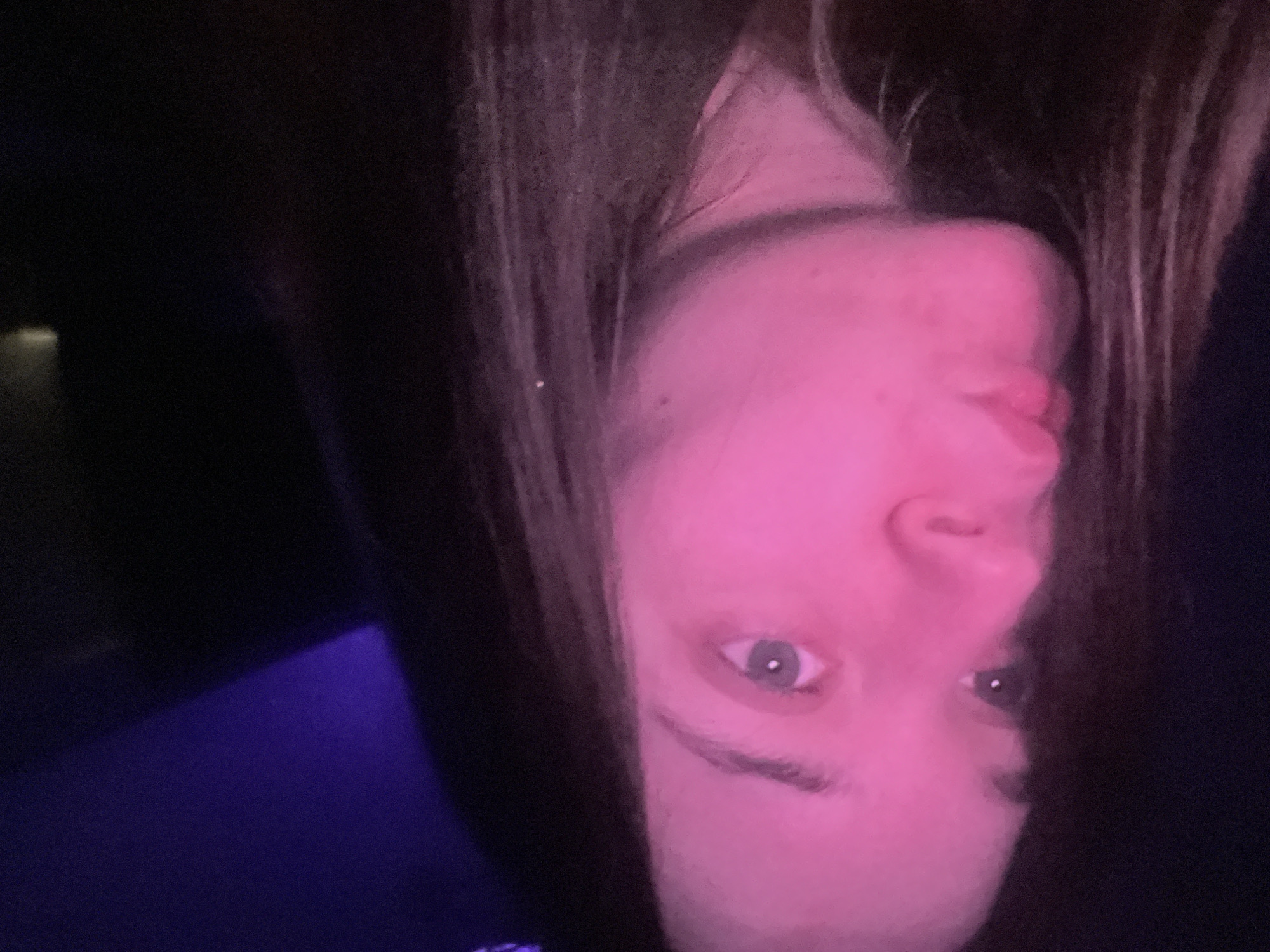 A female subject, appearing young, is depicted from the upper torso upwards, with her head turned to the viewer's left, and her gaze directed upwards and to the left. Her lips are pursed. The right side of her face (viewer's left) is intensely illuminated by a pink light source, casting a magenta hue across her skin. Her right eye (viewer's right) is visible, featuring a light-colored iris and a distinct pupil. Long, dark hair frames her face and extends downwards. The background is predominantly dark, with a distinct area of purple light visible in the upper right portion of the frame. The scene is located in the city of Pushchino, Russia.