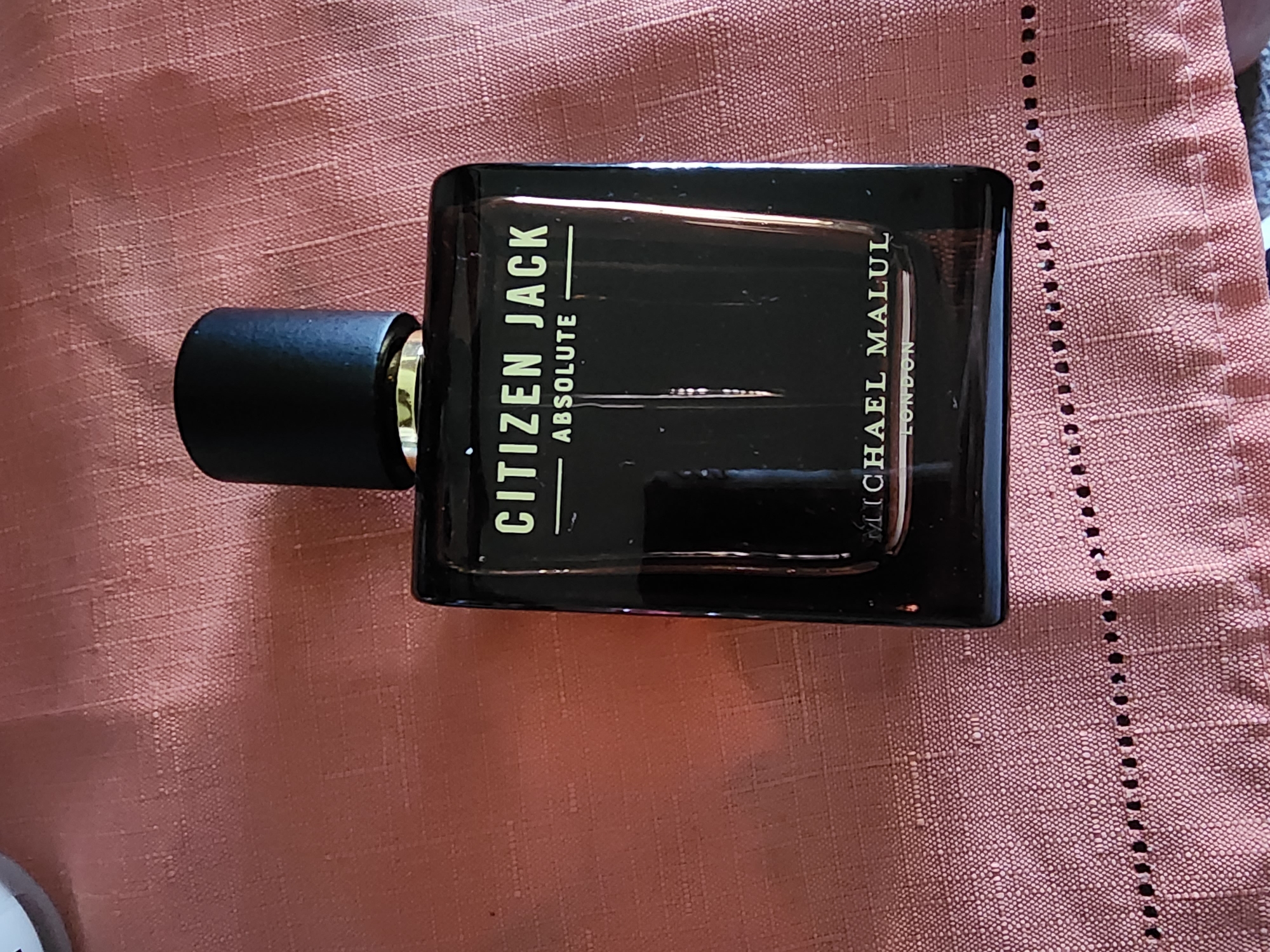 A black perfume bottle with a black cap sits on a textured pink tablecloth. The bottle has gold lettering that reads "CITIZEN JACK" and "ABSOLUTE" above "MICHAEL MALUL" and "LONDON." The background is out of focus. The image is taken from a slightly high angle.