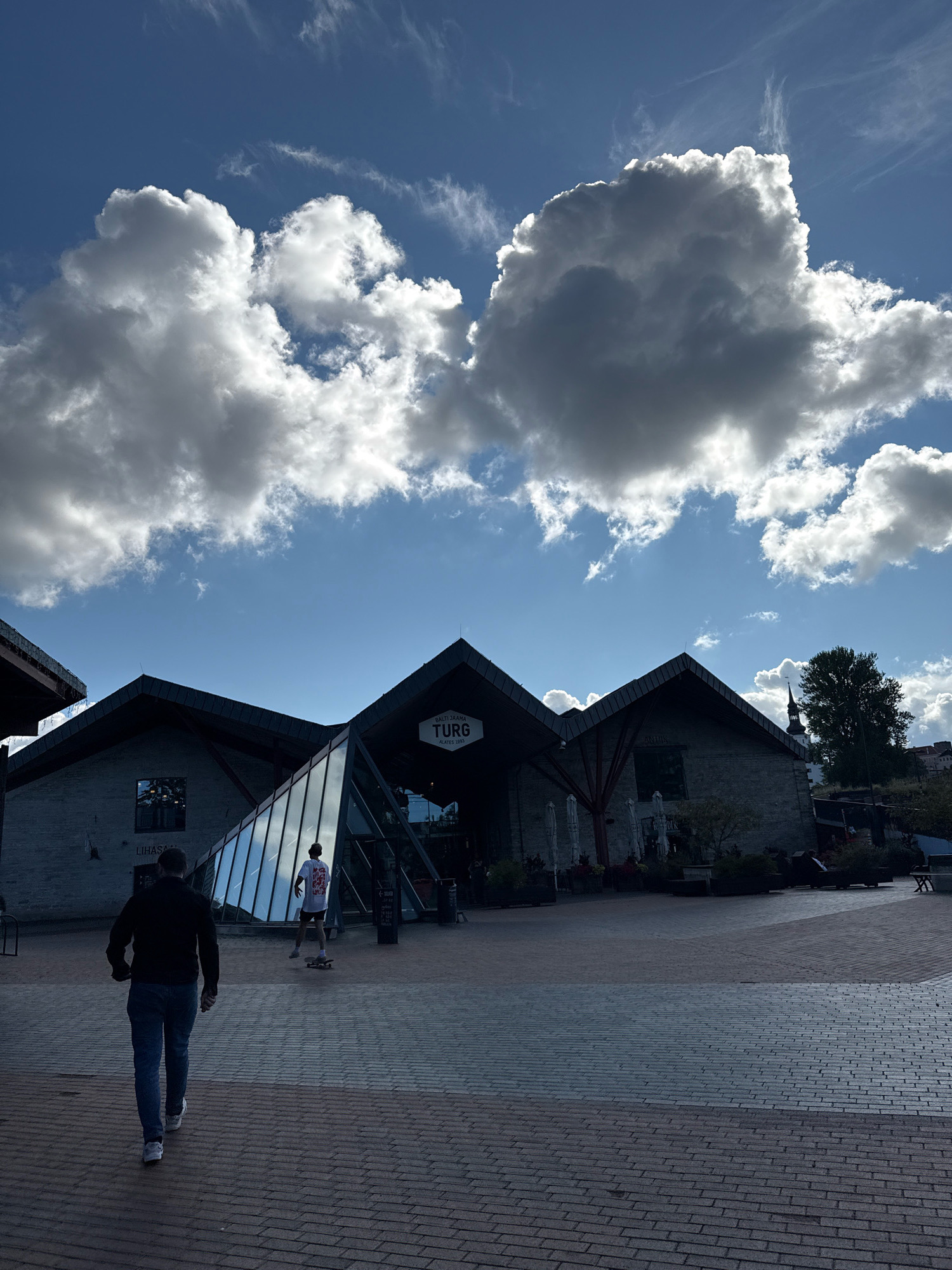 The image shows an exterior view of the Balti Jaama Turg in Tallinn, Estonia. The market hall, characterized by its dark, angular rooflines and brick exterior, is visible under a partly cloudy blue sky. In the foreground, a man in a dark jacket and blue jeans walks away from the camera, while another person on a skateboard is seen further ahead near a glass-paneled entrance to the building. The ground is paved with brickwork. The overall scene suggests an outdoor public space with a mix of architectural styles.