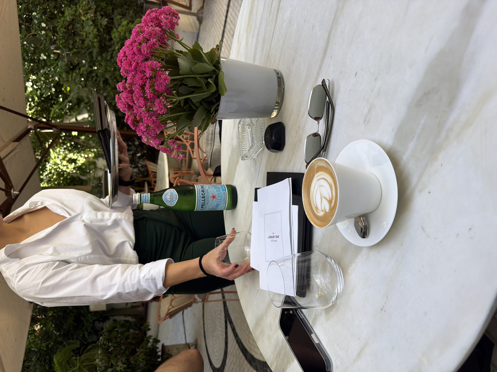 A waiter in a white shirt and green pants is serving a table at The Library Bar in Tel Aviv, Israel. The table is covered in a white marble surface. On the table, there is a white cup of coffee with latte art, a white saucer with a spoon, a pair of sunglasses, a glass of water, and a menu. A bottle of San Pellegrino water is also on the table, next to a bouquet of pink flowers in a white pot. A cell phone is also visible on the table. The scene appears to be an outdoor seating area with lush greenery in the background.