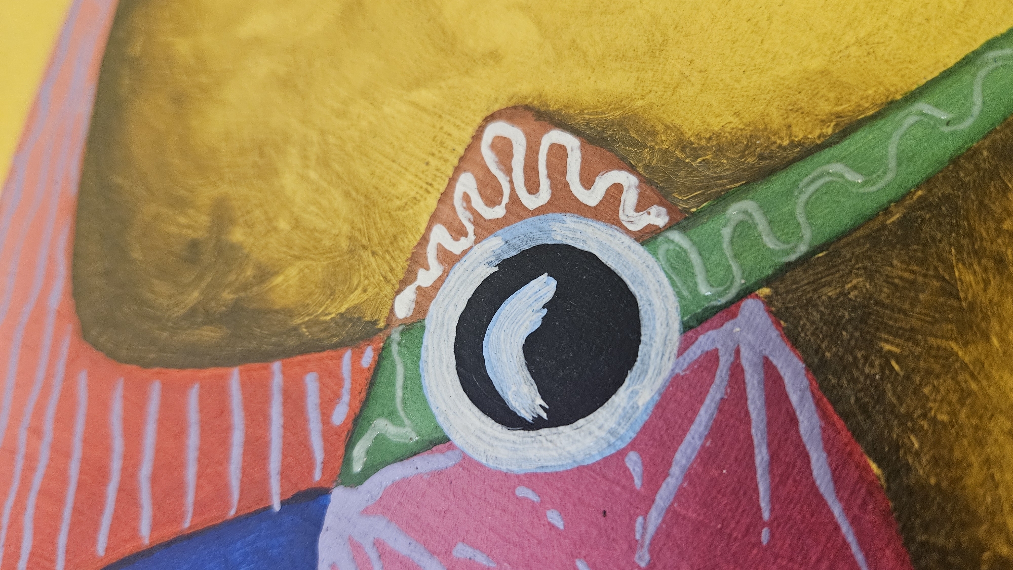 The image is a close-up, abstract painting of a fish or animal eye. The eye is black with a crescent-shaped blue pupil. It is surrounded by a white ring, then a light blue ring, and finally a dark blue outer ring. To the left of the eye is a pink area with purple lines radiating outwards. Above this is a section with horizontal red and white stripes. To the right of the eye is a green triangular shape with a white wavy line on it, leading to a rust-colored diamond shape also with a white wavy line. The background is a textured yellow and brown. The painting appears to be made with acrylics. There is no discernible location in Krasnodar, Russia, or any contextual information that would place it there.