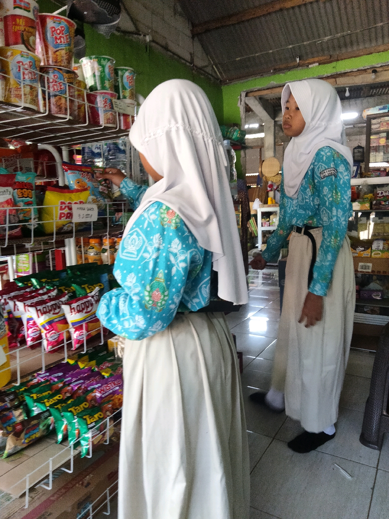In the city of Sidorejo, Indonesia, two young girls wearing white hijabs and long, light-colored skirts are browsing inside a small store. The girl on the left, seen from the back, wears a blue shirt with a white floral pattern. She is reaching for a snack on a shelf. The girl on the right, facing slightly away from the camera, wears a similar blue shirt and also has a white hijab. The store is filled with shelves packed with various food items, including packaged snacks, drinks, and instant noodle cups. The environment appears to be a modest, local shop with fluorescent lighting and a tiled floor.