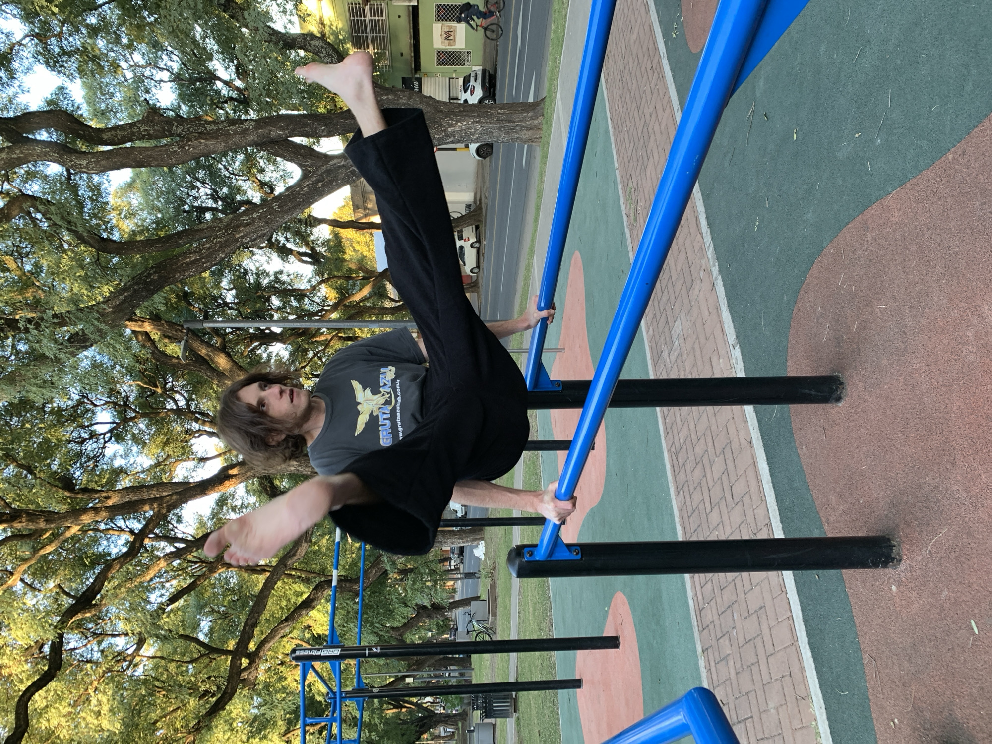A man is performing calisthenics on blue and black parallel bars in a park. He is upside down, with his feet pointing towards the top of the image and his hands gripping the bars. He is wearing a gray t-shirt and black pants, and is barefoot. The background features trees with dense foliage and a street with cars and pedestrians in the distance. The ground around the bars is covered with a textured surface, and a building is visible in the background. The location is a park in Buenos Aires, Argentina.