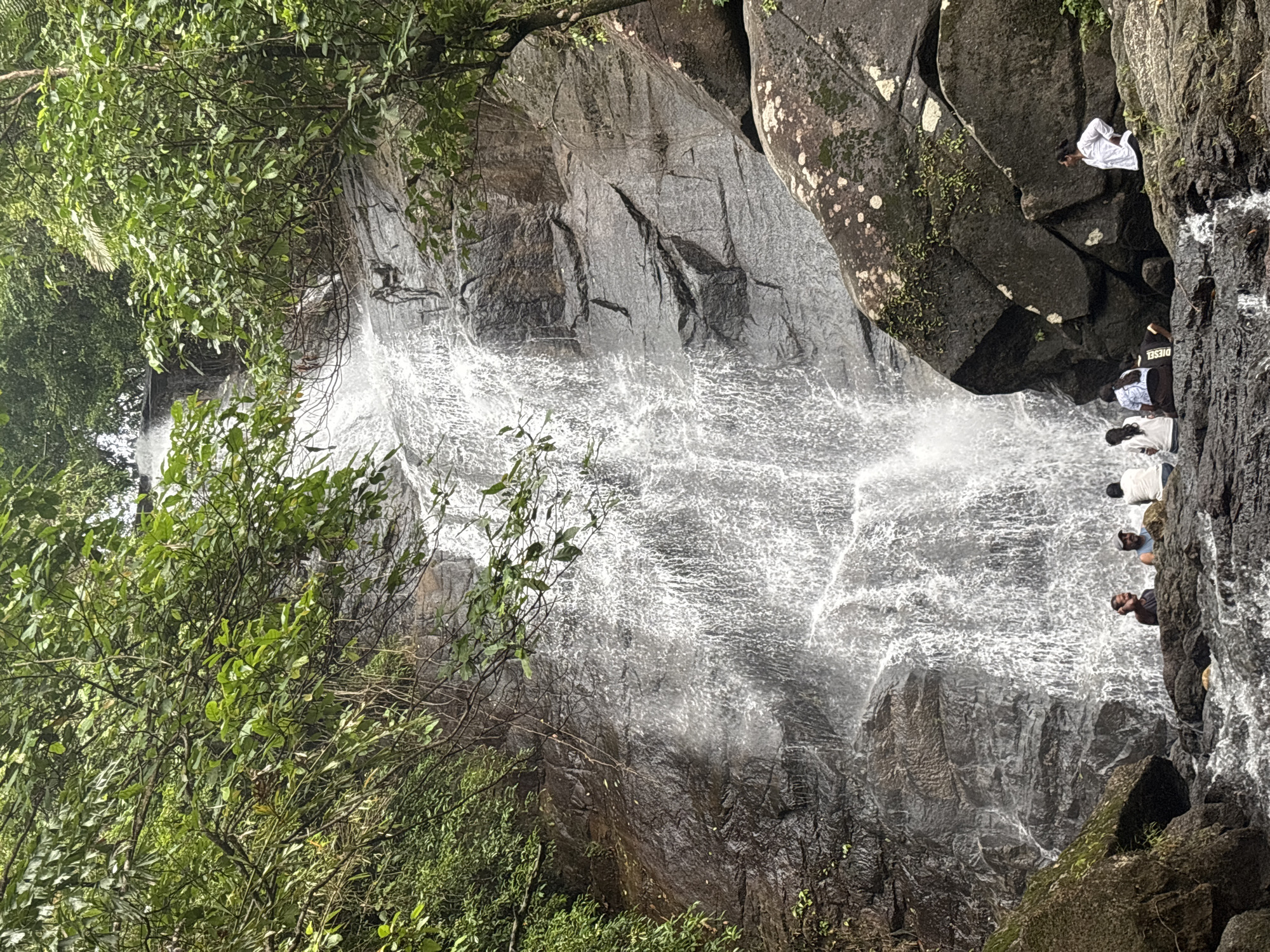 The image depicts a waterfall located in Ban Bang Khli, Thailand. The waterfall cascades down a rocky cliff face, with lush green vegetation visible on either side. Several people are positioned near the top of the waterfall, standing or sitting on rocks near the water's edge. The scene is captured in daylight, suggesting outdoor conditions.