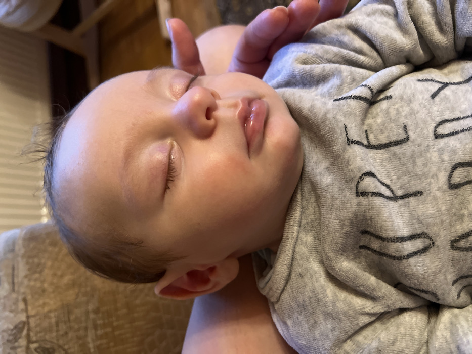 A baby sleeps peacefully, resting on someone's lap. The child's face is turned towards the viewer, eyes closed, with a hand gently touching their forehead. The skin is smooth and soft, with a rosy tint to the cheeks. They are wearing a gray onesie with a pattern of black letters. The background is blurred, but appears to be a warm, indoor setting.