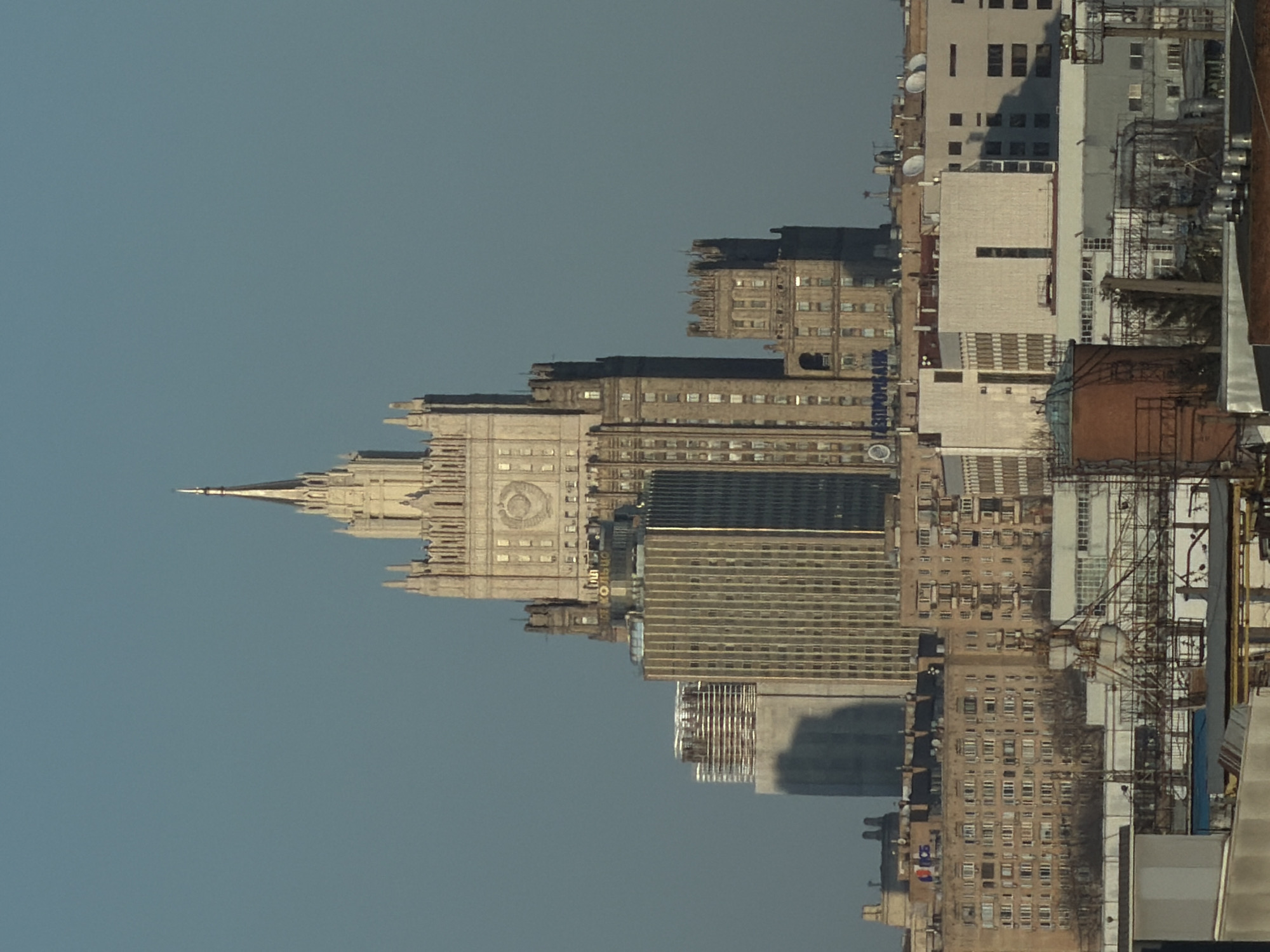 The image shows a cityscape in Moscow, Russia. The most prominent building is the Ministry of Foreign Affairs, one of the Seven Sisters skyscrapers built in the Stalinist architectural style. The building has a distinct spire and clock on its facade. Other buildings of varying heights and architectural styles are visible, with some having satellite dishes on their roofs. Construction or maintenance infrastructure is present. The sky is a clear blue.