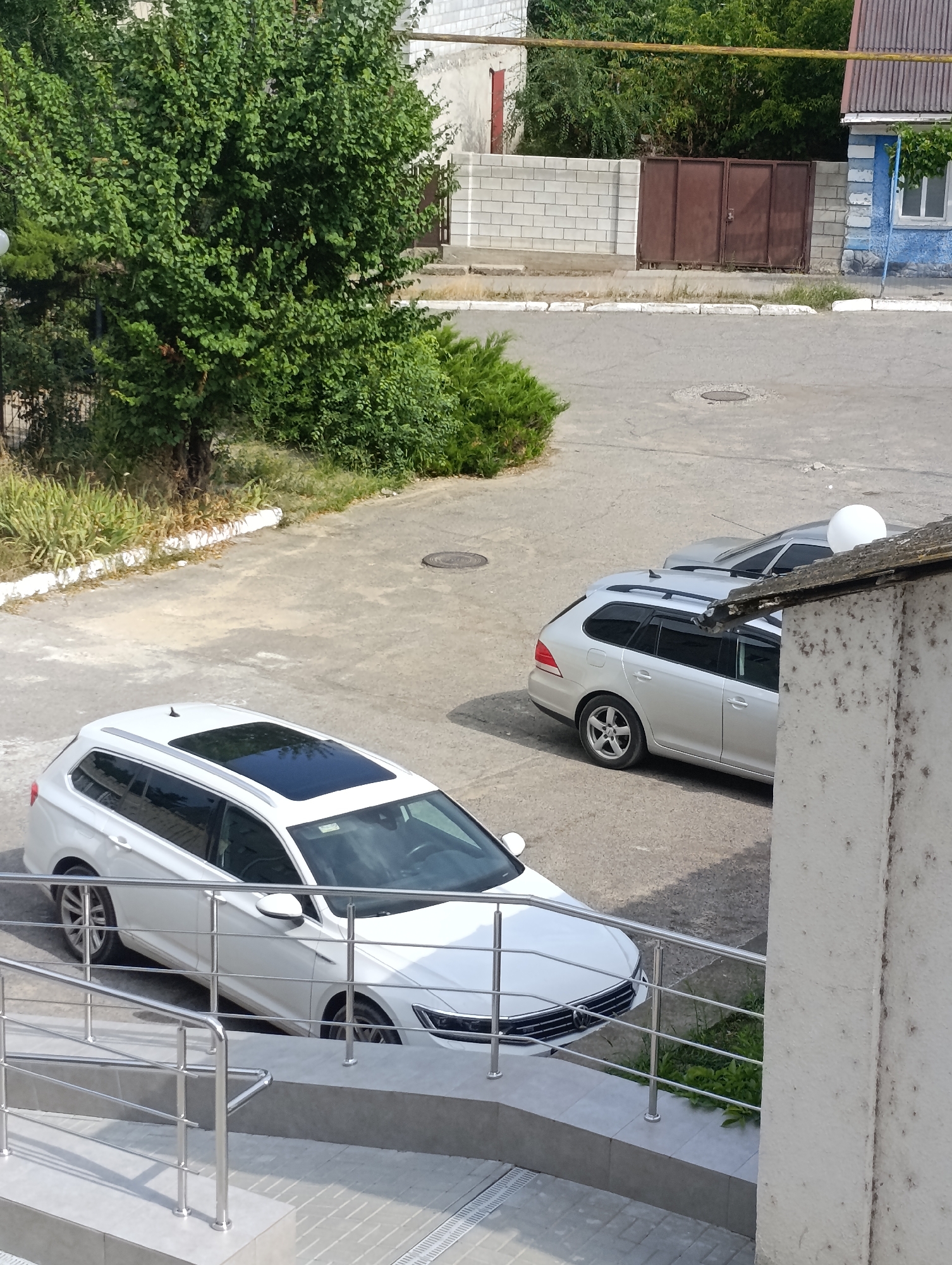 In Dubăsari town council, Moldova, a sunlit daytime scene unfolds in an urban environment. Two cars, a white station wagon with a sunroof and a silver station wagon, are parked on a paved area. The white car is positioned in the foreground, angled towards the left, revealing its front grille and headlights. The silver car is parked further back, to the right of the white car, its side profile visible. A white satellite dish is mounted on the roof of a building to the right of the silver car. The background features a white brick wall, a brown metal gate, a blue building with a window, and lush green trees. The ground is asphalt, with some manhole covers visible. A metal railing lines the edge of an elevated walkway in the foreground, leading towards the white car.