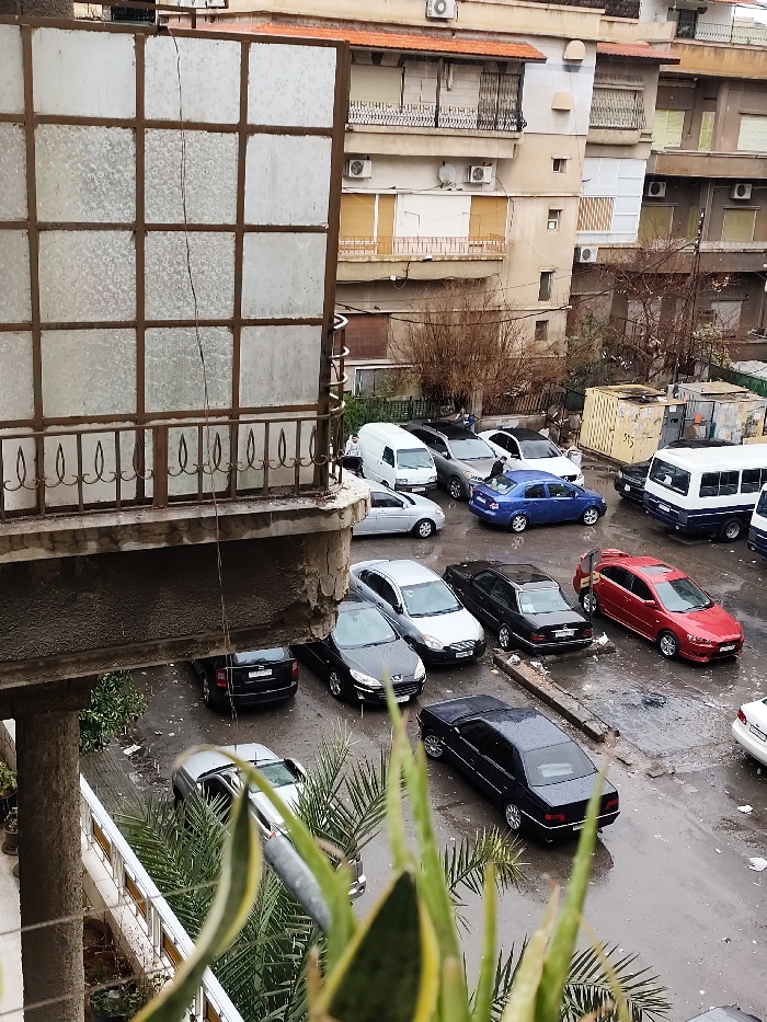 The image depicts an elevated view of an outdoor parking area within the city of As-Salihiya Municipality, Syria. The scene appears wet, suggesting recent rainfall.
In the immediate foreground, from a balcony perspective, a large multi-paned structure with frosted or textured glass and a dark frame partially obscures the upper left view. A thin, dark cable or wire runs vertically down its center. Below this, a concrete balcony edge exhibits chipped and deteriorated sections, with a decorative dark metal railing visible. Green foliage, likely Sansevieria and palm fronds, is present at the bottom of the frame, originating from below the balcony.
The midground is dominated by a wet, asphalt or concrete parking lot containing numerous parked vehicles. These include a white utility van, a silver SUV, a white sedan, a blue sedan, a black sedan, a red sedan, a dark blue and white mini-bus, and several other black and silver sedans and hatchbacks. One individual is observed standing near the rear of the blue sedan, appearing to interact with the vehicle. Another figure is faintly visible near the silver SUV. The ground throughout the parking area is visibly wet with scattered puddles. A broken concrete curb section lies on the ground near the center-right. A yellow corrugated metal container or skip is positioned at the far right of the parking area.
In the background, multi-story residential buildings rise, featuring balconies with metal railings, various windows (some with closed roller shutters or blinds), and multiple external air conditioning units. Sparse, bare-branched trees or large shrubs are situated behind the parking lot. The sky overhead is overcast, providing diffuse lighting to the scene.